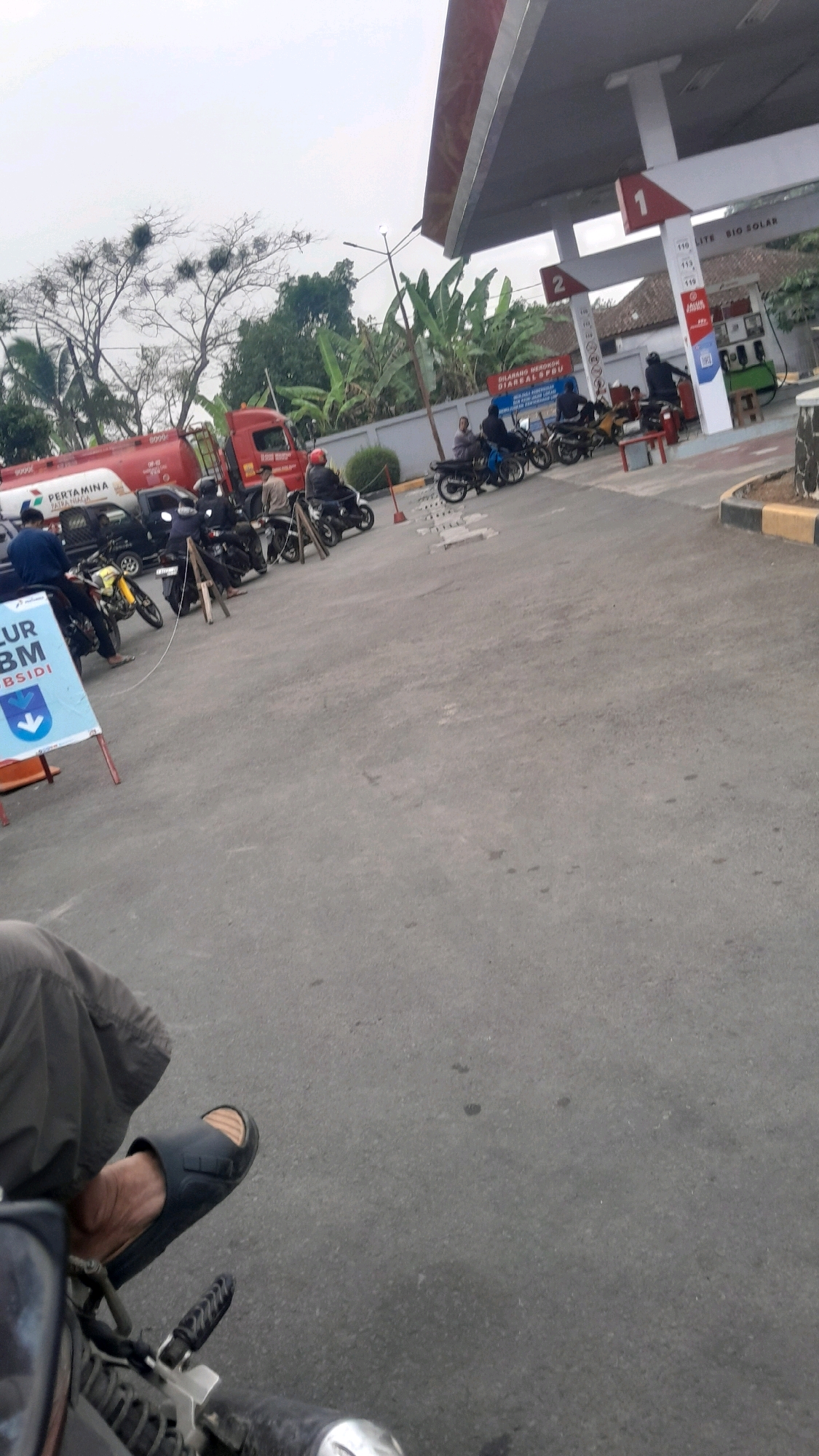 The image depicts a Pertamina gas station in Subang, Indonesia. In the foreground, a person wearing gray pants and a black sandal is seated on a motorcycle. The motorcycle's handlebar and part of its body are visible. The background shows a red Pertamina fuel tanker truck. Several individuals on motorcycles are lined up near the gas pumps under a covered canopy. One motorcycle is yellow, while others are black. A sign on the left side of the image reads "LUR BBM SUBSIDI" in blue and white. The environment appears to be an outdoor paved area with trees and vegetation visible in the distance. The sky is overcast.