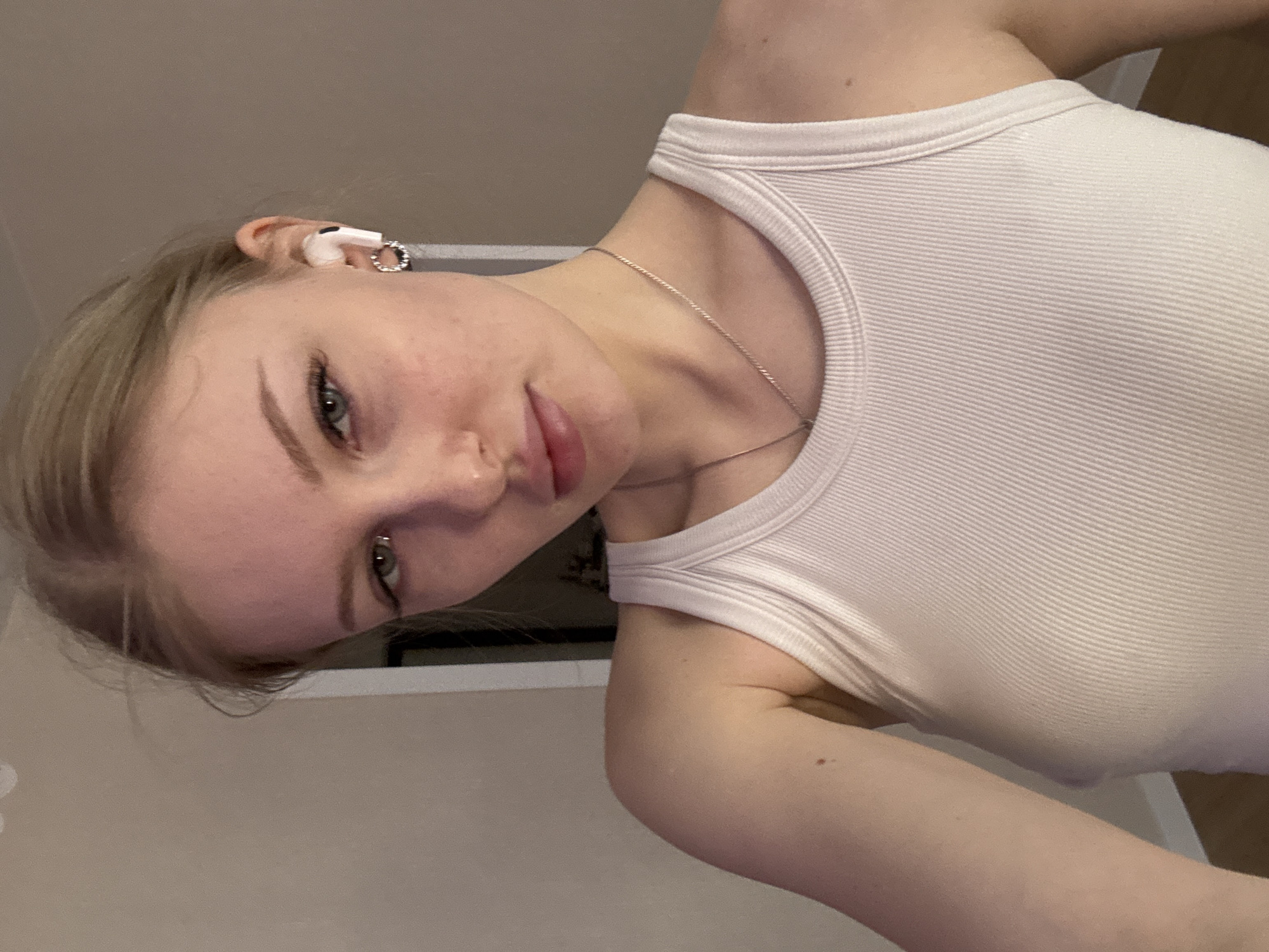 A young woman with fair skin and blonde hair, wearing a white ribbed tank top and a silver necklace, looks at the camera. She has an earbud in her right ear and an earring in the same ear. Her head is slightly tilted to the left. The background appears to be a plain wall in an interior space. There is no indication of the location being Perm, Russia.