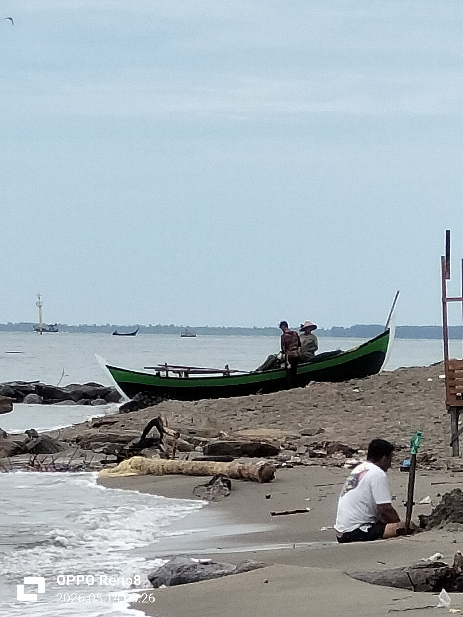 The photograph depicts a coastal scene, likely in Lhokseumawe, Indonesia. In the foreground, a man wearing a white t-shirt and dark shorts sits on the sandy beach near the shoreline, facing away from the camera. Beside him is a pole stuck into the sand.
Further back on the beach, a dark green and black boat rests on the sand, appearing to be a traditional fishing vessel. Two figures are sitting inside the boat, partially obscured, one wearing a wide-brimmed hat.
The beach consists of dark-colored sand littered with rocks and driftwood. Small waves are lapping against the shoreline. In the background, the ocean extends to the horizon under a cloudy sky. A structure resembling a lighthouse or navigational aid is visible in the distance. Several small boats are also visible in the background.
A wooden structure stands on the right side of the image.