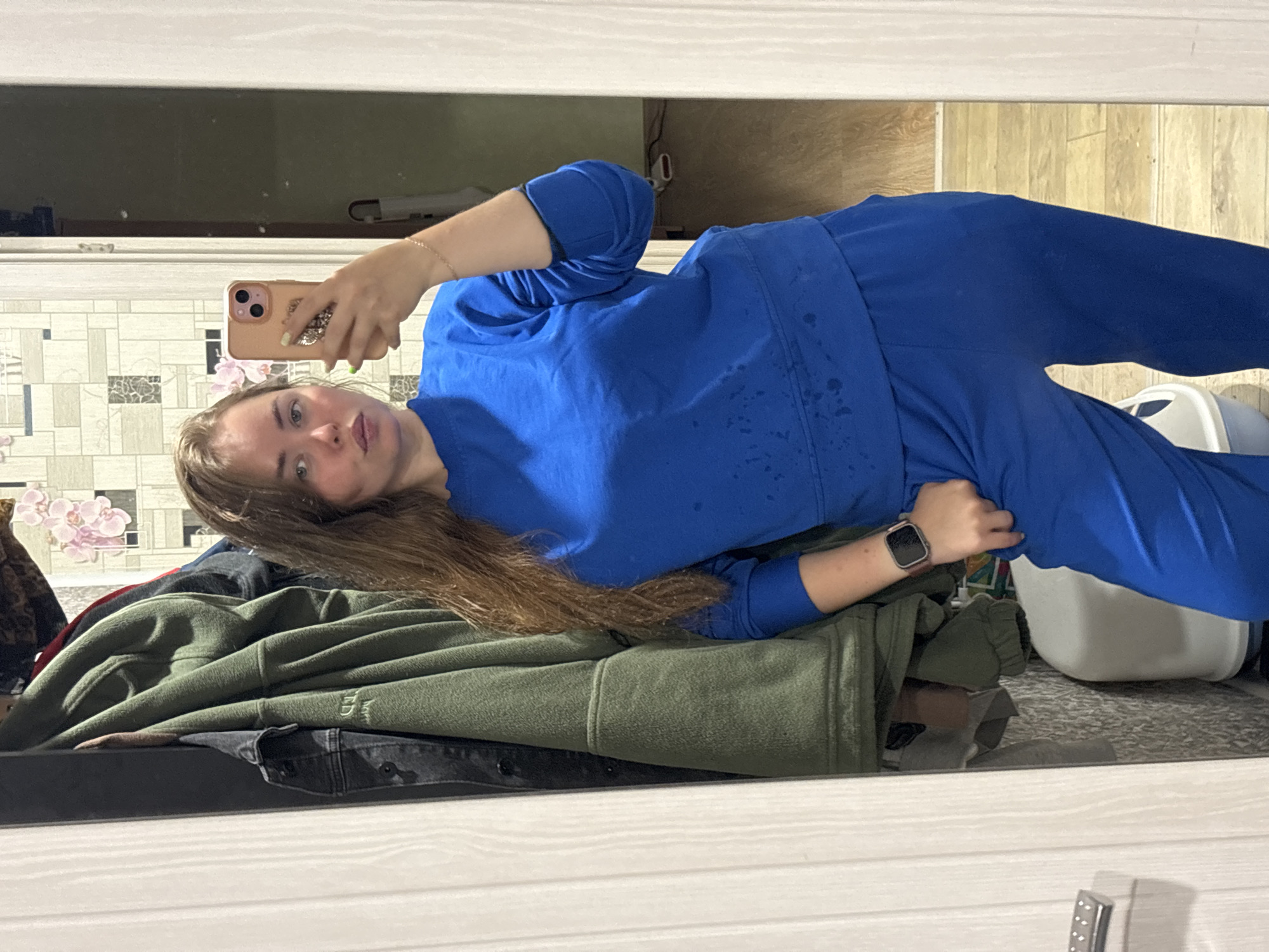 A young woman in a blue tracksuit is taking a selfie in a mirror. She is lying on a surface covered with clothes, including a green sweatshirt and a black jacket. Behind her is a wall with a patterned wallpaper featuring floral designs. The floor is wooden, and a white object resembling a cat litter box is visible. The location is implied to be Izhevsk, Russia, but no specific landmarks or details confirm this.