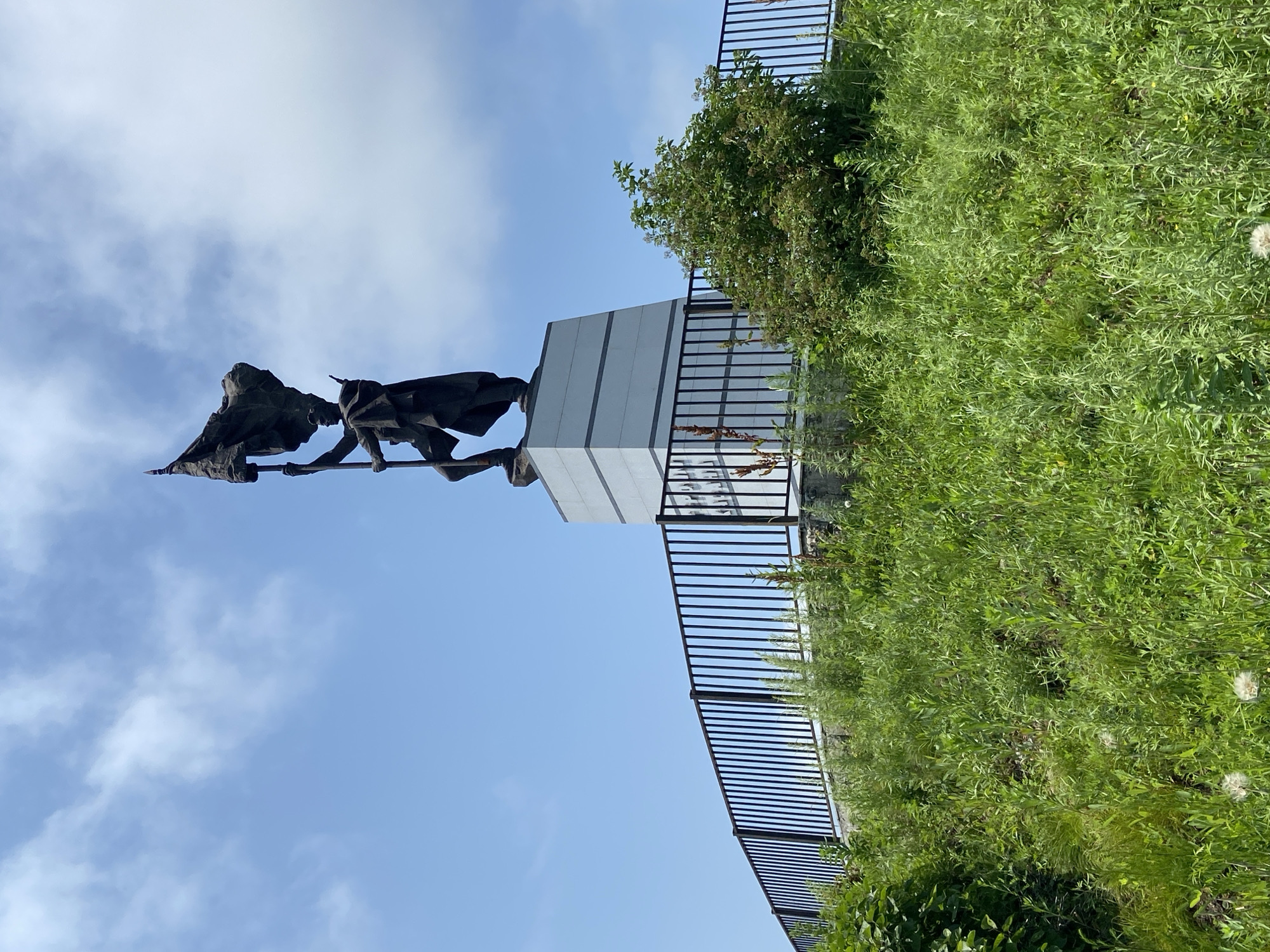 A statue of a soldier holding a flag aloft stands on a white pedestal, which is surrounded by a black metal fence. The statue and pedestal are situated on a grassy hill in Kraskino, Russia. The sky is blue with scattered white clouds. The hill is covered in green grass and some wildflowers.