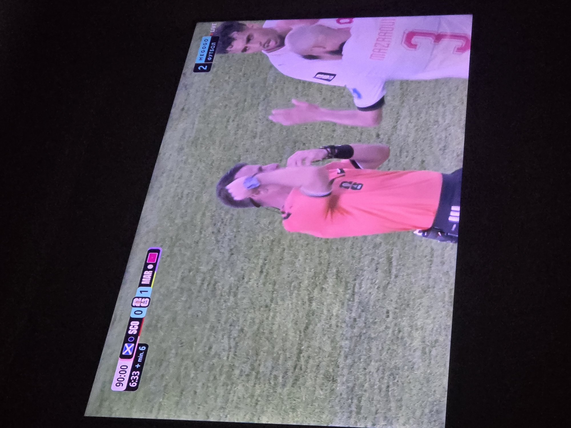 A television screen displays a soccer match. The score is displayed as 0-1 in favor of "MAR". The time is 90:00, with an additional 6 minutes indicated. A referee in an orange jersey is interacting with a player in a white jersey who has the name "MAZARROUI" and the number "3" on his back. The referee appears to be speaking to the player. The environment is a soccer field with green grass. The screen also shows logos for "MEGOGO" and "ФУТБОЛ", indicating the broadcast platform and language. The scene is viewed from a slight angle, suggesting it is being watched on a screen in a room.