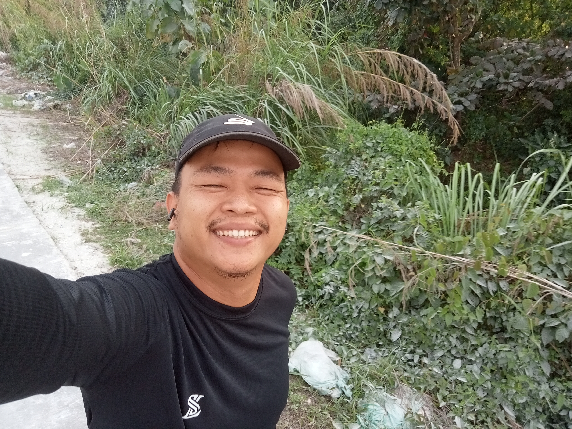 A man, wearing a black t-shirt and a black baseball cap, smiles broadly at the camera. He has earbuds in both ears and appears to be taking a selfie. The man is standing outdoors, with lush green foliage and tall grasses surrounding him. In the background, a dirt path can be seen, and a few white plastic bags are scattered on the ground. The lighting suggests it is daytime. The image is consistent with a location in the city of Teluk Kenidai, Indonesia.