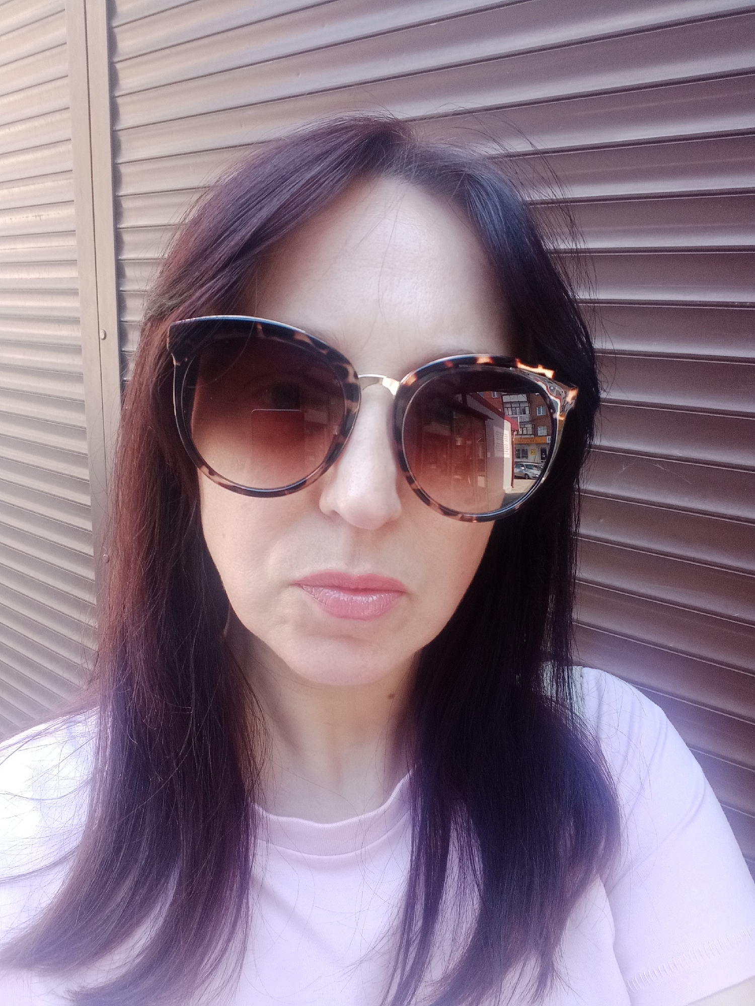 A woman with long, dark hair and sunglasses is shown in a medium shot, facing forward against a dark, ribbed background. She is wearing a light pink t-shirt. The sunglasses have a tortoiseshell pattern and reflect buildings and a car. The background appears to be an exterior shutter or garage door.