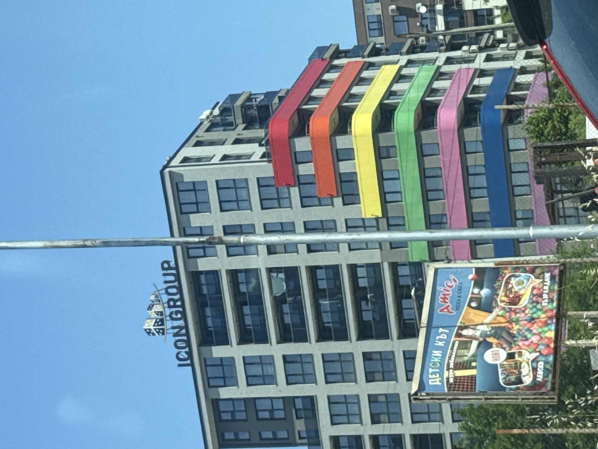 This is an outdoor, eye-level shot of a modern apartment building in the city of Varna, Bulgaria, with a billboard in the foreground. The building is tall and grey, with many windows and balconies. Some of the balconies have colorful, curved edges, resembling a rainbow. The billboard, on the left side of the image, features an advertisement for a children's play area and a pizza and grill restaurant called "Amica's". The billboard shows children playing in a ball pit and images of the restaurant. The sky is clear and blue. A metal pole, possibly a street lamp, runs vertically through the right side of the frame.