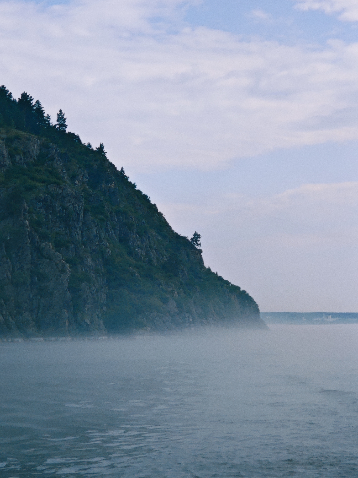 The image shows a steep, rocky cliff covered in dark green vegetation and scattered pine trees. The cliff slopes down towards a body of water, which is partially obscured by a thick layer of fog or mist. The water surface shows small ripples. In the distance, a hazy horizon line is visible, suggesting the presence of land or structures shrouded in mist. The sky above is a pale blue with scattered, wispy white clouds. The overall atmosphere is muted and somewhat moody due to the fog and overcast sky. The scene is a natural landscape with no visible people or human activity.