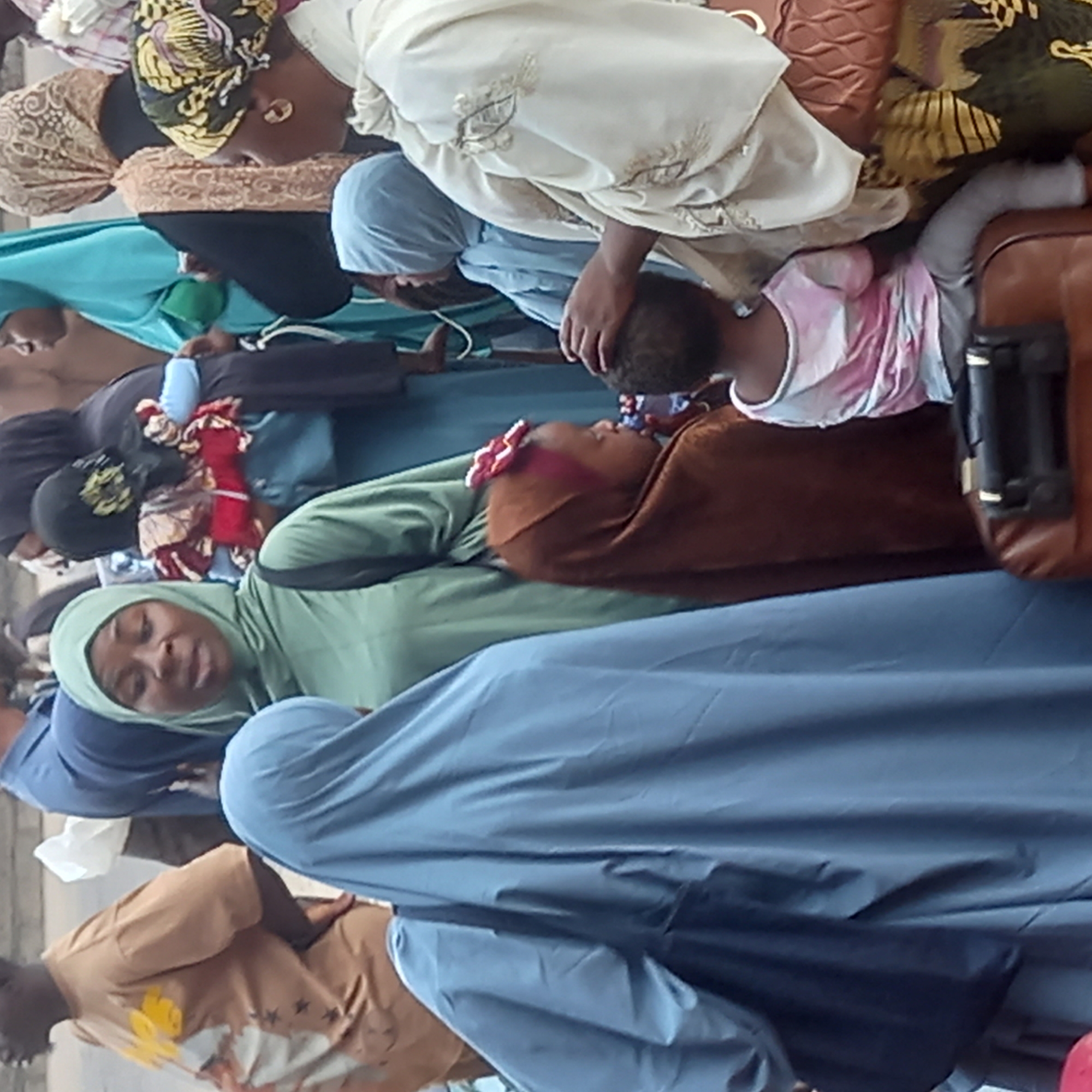 In Sardauna, Nigeria, a group of women wearing hijabs and other traditional clothing are gathered together. One woman is holding a small child. Luggage and other personal belongings are nearby. The environment appears to be outdoors with a grey or light-colored ground surface.