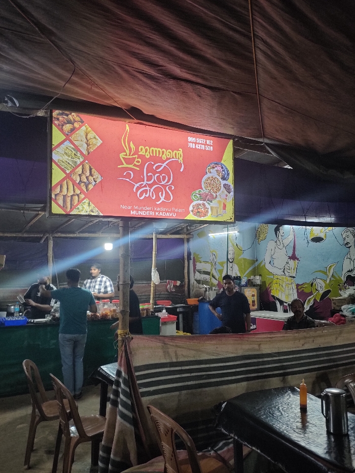 The image captures a bustling, casual food stall or restaurant at night in Munderi, India. The setting appears to be semi-open-air, characterized by a dark tarpaulin ceiling supported by bamboo poles, giving it a rustic or temporary feel.
Central to the scene is a large, illuminated red and yellow sign featuring Malayalam script and images of food. The Malayalam text reads "മുന്നൻ്റെ കലം" (Munnan'te Kalam), translating to "Munnan's Pot" or "Munnan's Vessel." Below this, English text indicates the location: "Near Munderi kadavu Palam, MUNDERI KADAVU." Two phone numbers, 989 5672 102 and 790 4378 539, are also visible. The sign displays photographs of various traditional Indian snacks, such as fried fritters and sweet pastries, on its left side, and plates of full meals or curries on its right.
Several men are present, suggesting ongoing operations. Two men are busy behind a counter on the left, one facing away in a dark green t-shirt and jeans, and another in a plaid shirt. They appear to be involved in food preparation or serving, with food containers visible on the counter. Further to the right, another man in a black t-shirt leans against a different counter. A fourth man, with a darker complexion, is seated at a table on the far right.
The back wall features a colorful mural depicting figures in what appears to be traditional or rural attire, possibly engaged in farming or local activities, adding a cultural touch to the establishment. The lighting is artificial, with a bright overhead light illuminating the counter area and the glow from the sign. The scene indicates nighttime, as the surroundings beyond the stall's lights are dark. Empty plastic chairs and tables are arranged for customers, though the tables in the foreground show only a sauce bottle and a steel container on one. The overall ambiance is that of a local, unpretentious eatery serving a variety of dishes and snacks.