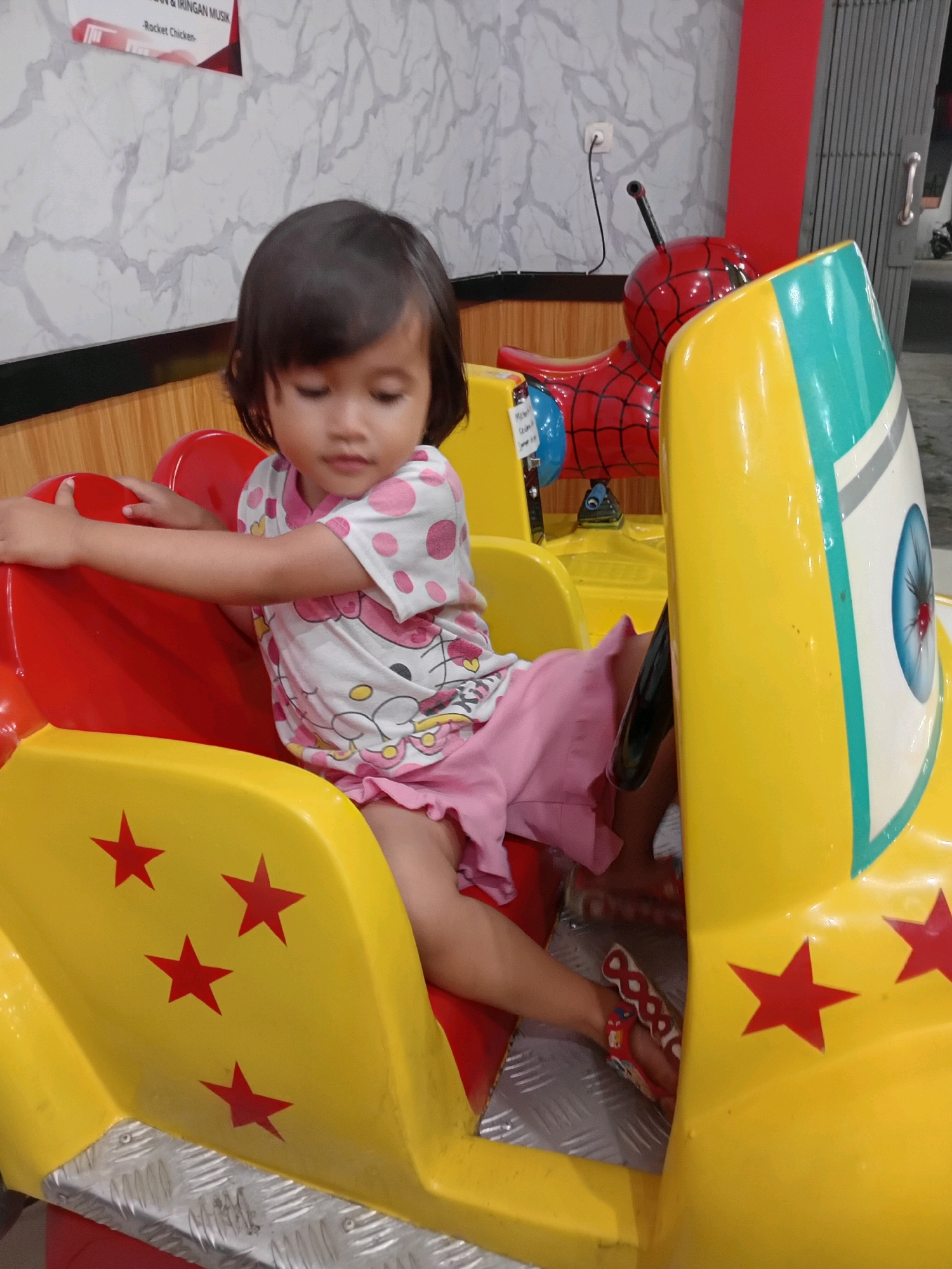 A young girl with dark hair and a pink and white polka dot shirt sits in a yellow and red amusement ride designed to resemble a car. She is wearing pink shorts and red sandals. The ride features red stars and a metallic floor. In the background, a Spiderman-themed amusement ride is visible. The scene is illuminated by artificial light.