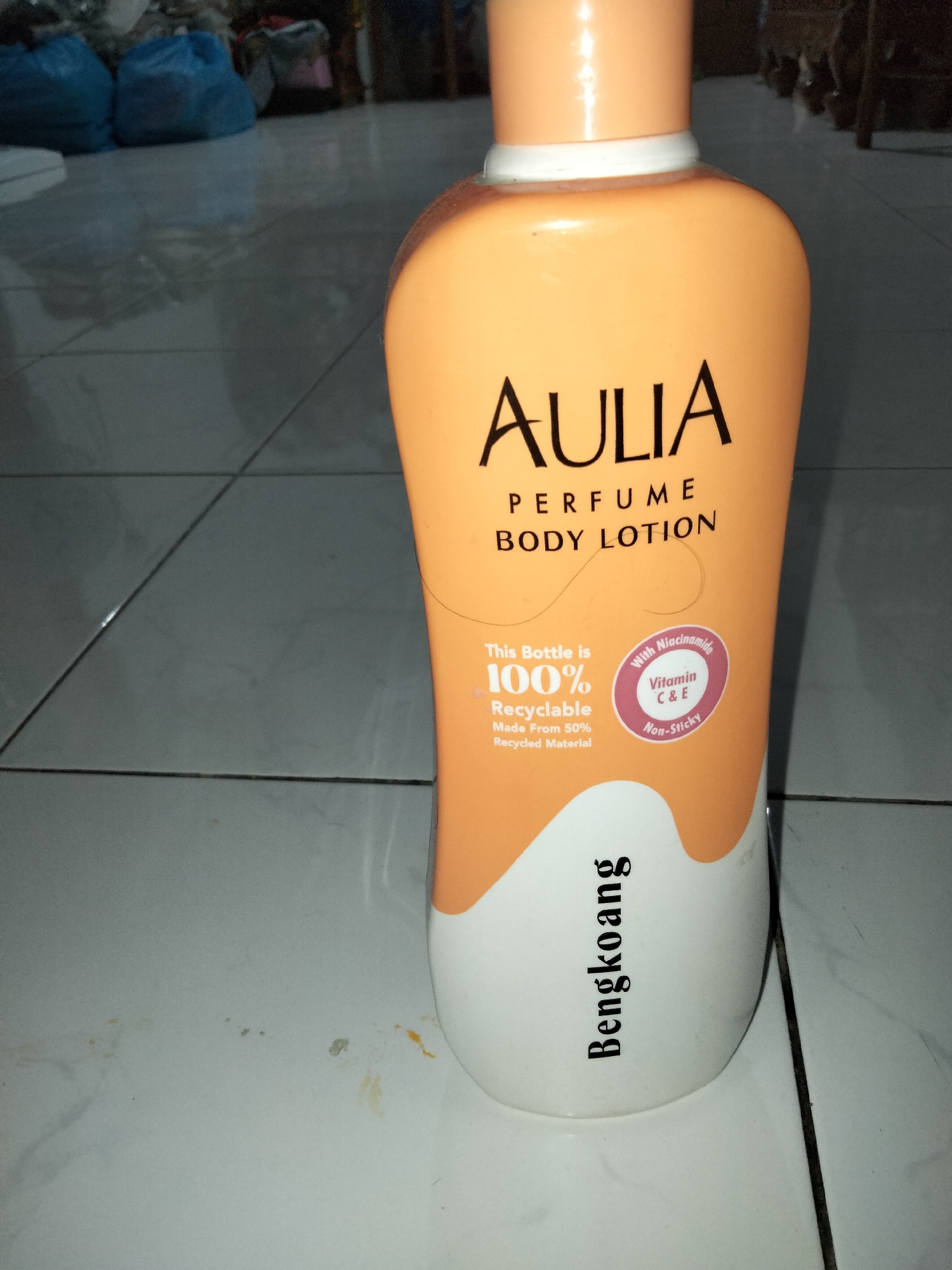 In Payagaboh, Indonesia, a partially visible orange and white plastic bottle of Aulia Perfume Body Lotion is positioned on a tiled floor. The bottle, standing upright, features prominent text indicating "AULIA PERFUME BODY LOTION". A circular graphic on the bottle states "This Bottle is 100% Recyclable Made From 50% Recycled Material" and also mentions "With Niacinamide Vitamin C & E Non-Sticky". The word "Bengkoang" is printed vertically on the lower white portion of the bottle. The background consists of a tiled floor with visible grout lines and a blurred reflection of items in the upper left corner, possibly plastic bags.
