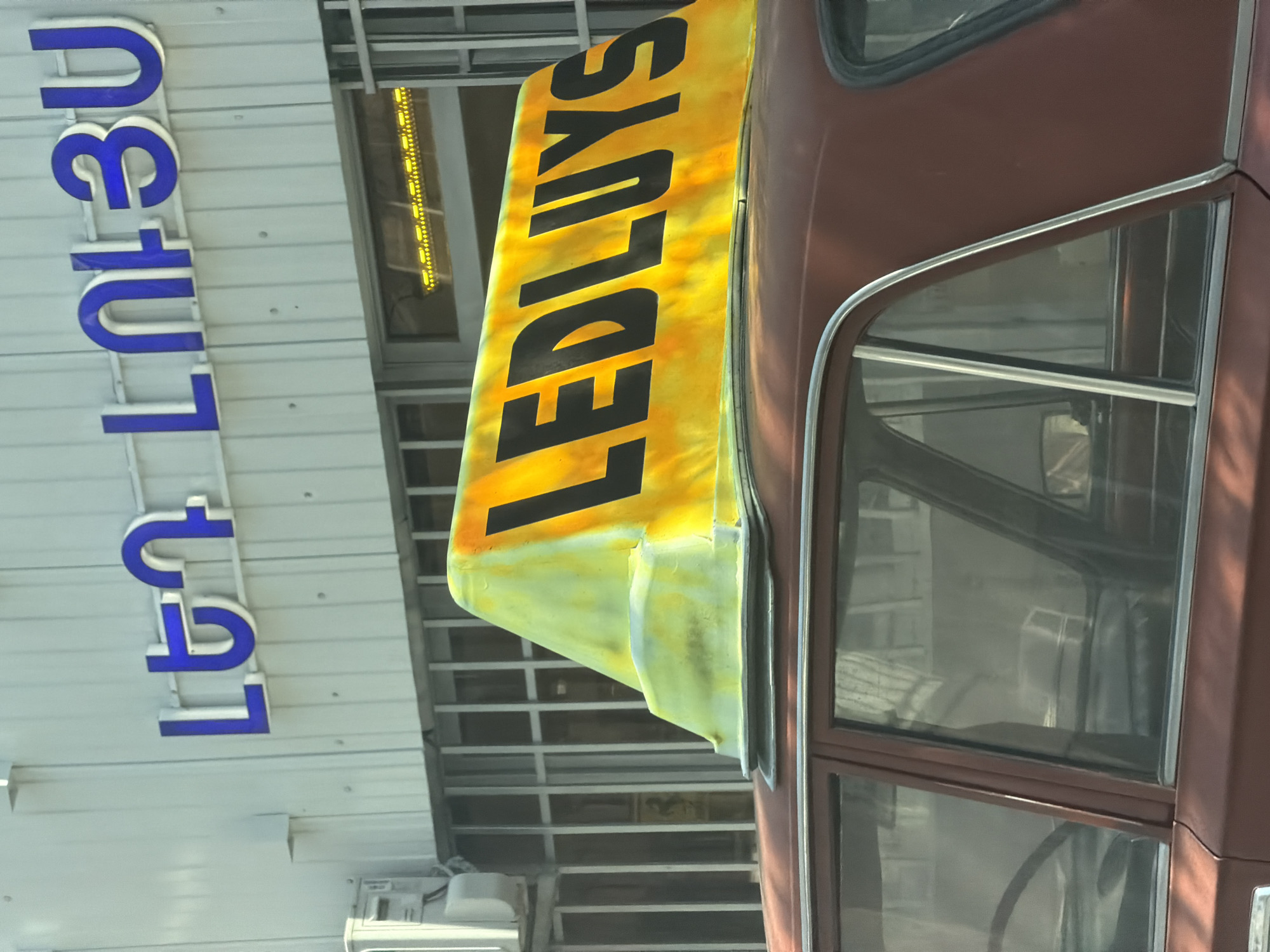 The image shows a red Soviet-era car with a yellow taxi sign on its roof. The sign displays the word "LEDLUYS" in black letters. The car is parked in front of a building with a white facade and blue lettering, which reads "ԼԵՆ ԼՈՒՅՍ". The blue lettering is in Armenian. This translates to "LEN LUYS", which is a taxi company in Yerevan, Armenia. The building appears to be a commercial establishment, with barred windows and what looks like an air conditioning unit on the left side. The sunlight casts a warm glow on the scene.