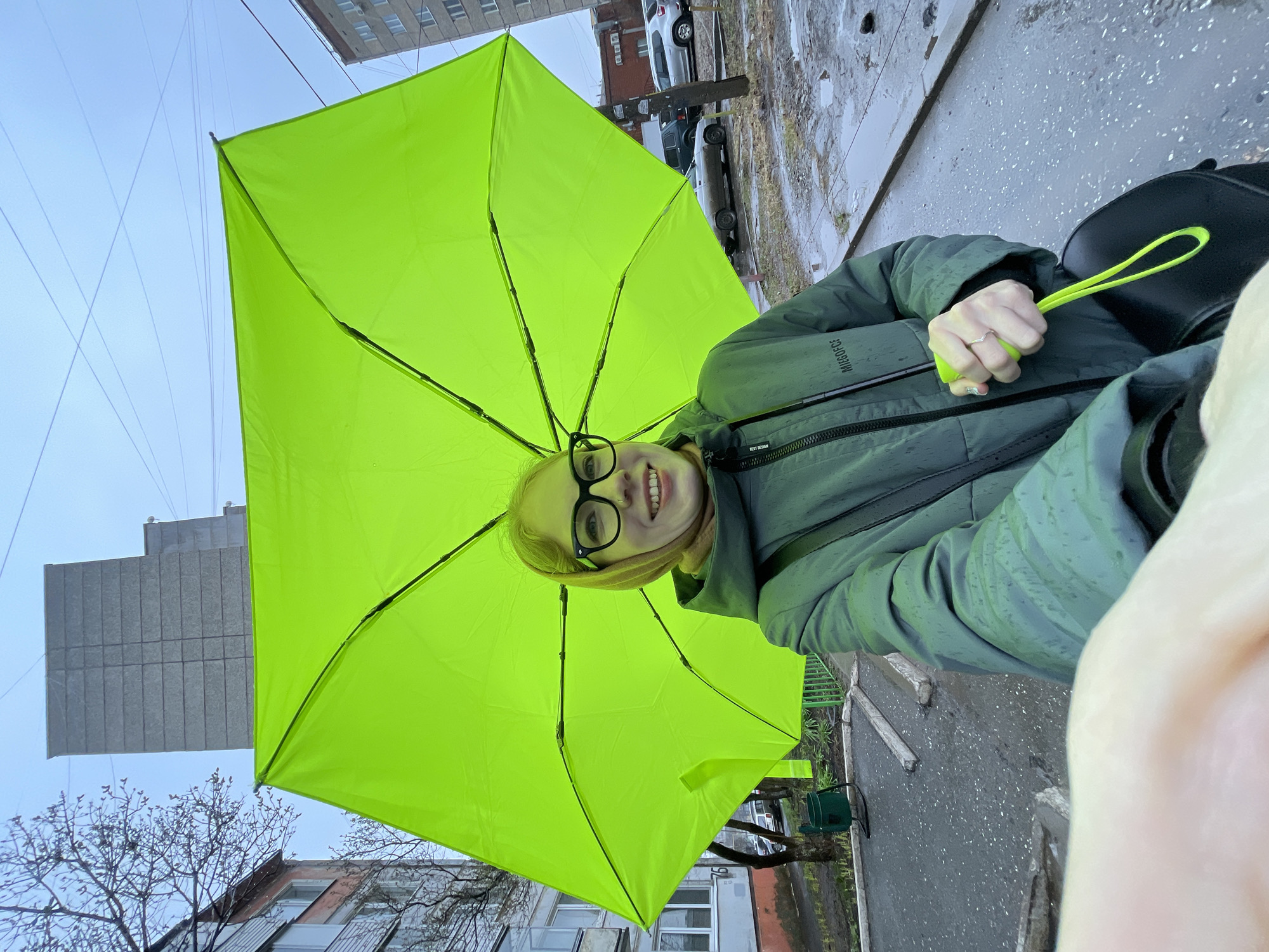 The image captures a smiling young woman holding a vibrant lime green umbrella in an urban setting in Izhevsk, Russia. The photo is taken from a low angle, looking up at her face and the open umbrella, which dominates the upper portion of the frame.
The woman has light-colored hair and wears black-rimmed glasses, looking directly at the camera with a cheerful expression. She is dressed in a dark teal or greyish-green hooded jacket, with a lighter scarf visible around her neck. Her left hand, adorned with a silver ring, holds the umbrella's handle, which features a matching neon green lanyard. Visible text on her jacket includes "MIESOFCE" on the upper sleeve and "BEST DESIGN" closer to the cuff.
The background reveals a wet, asphalt street or sidewalk with visible puddles, indicating recent rain or damp weather. Several parked cars of various models, including a white sedan and a darker SUV, line the street. Tall, multi-story residential buildings with numerous windows rise in the background, characteristic of urban architecture. Bare trees suggest a cooler season, possibly late autumn or early spring. The sky appears overcast. The overall impression is a moment of cheerfulness amidst a somewhat gray, rainy day.