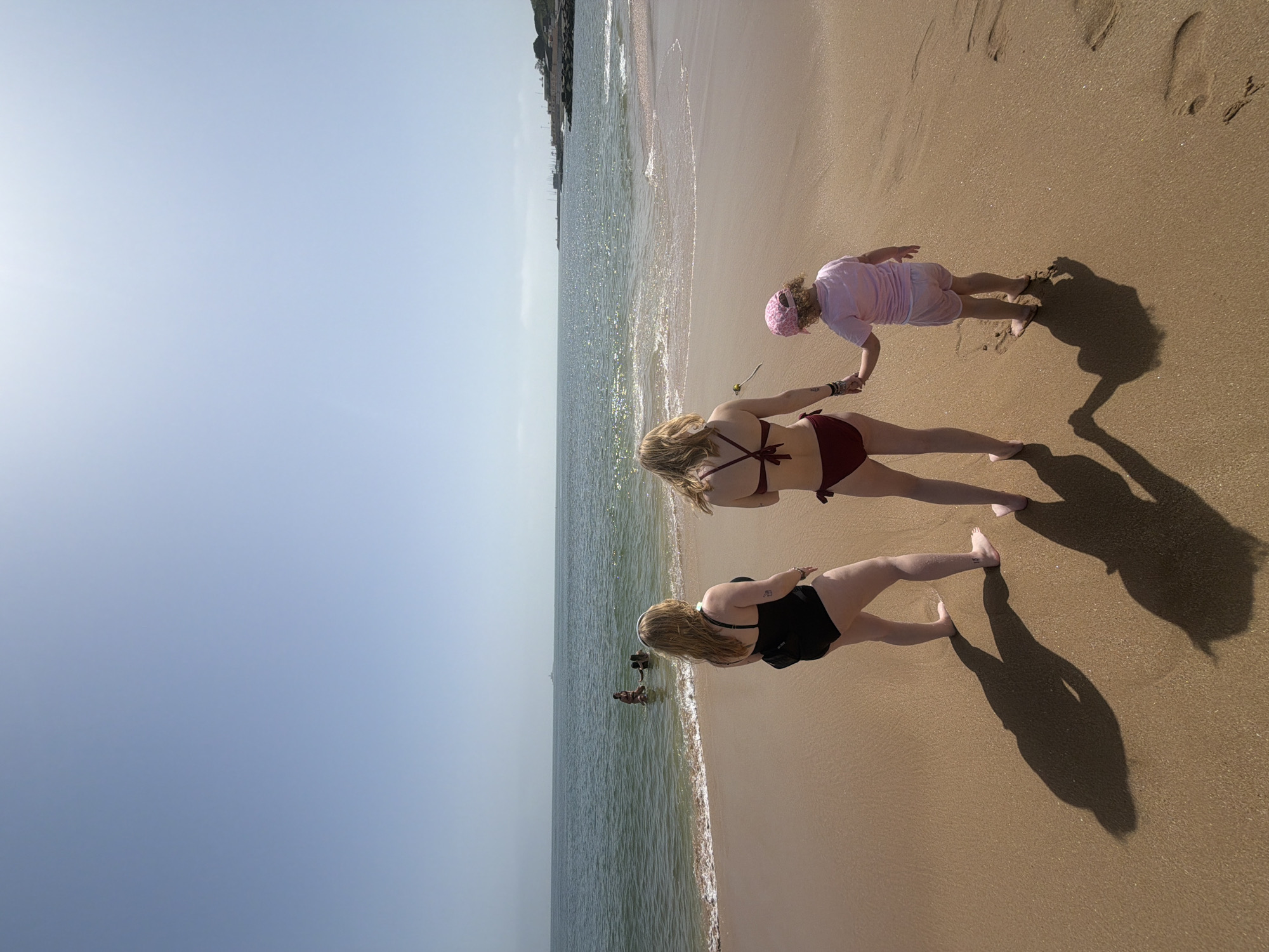 The scene depicts three females standing on a sandy beach next to the ocean. A child wearing a pink outfit is being held by a blonde female wearing a maroon bikini. Another female with blonde hair is next to them, wearing a black one-piece swimsuit. The sun is bright and the shadows are dark. There are people in the ocean. The beach is in Oeiras, Portugal.