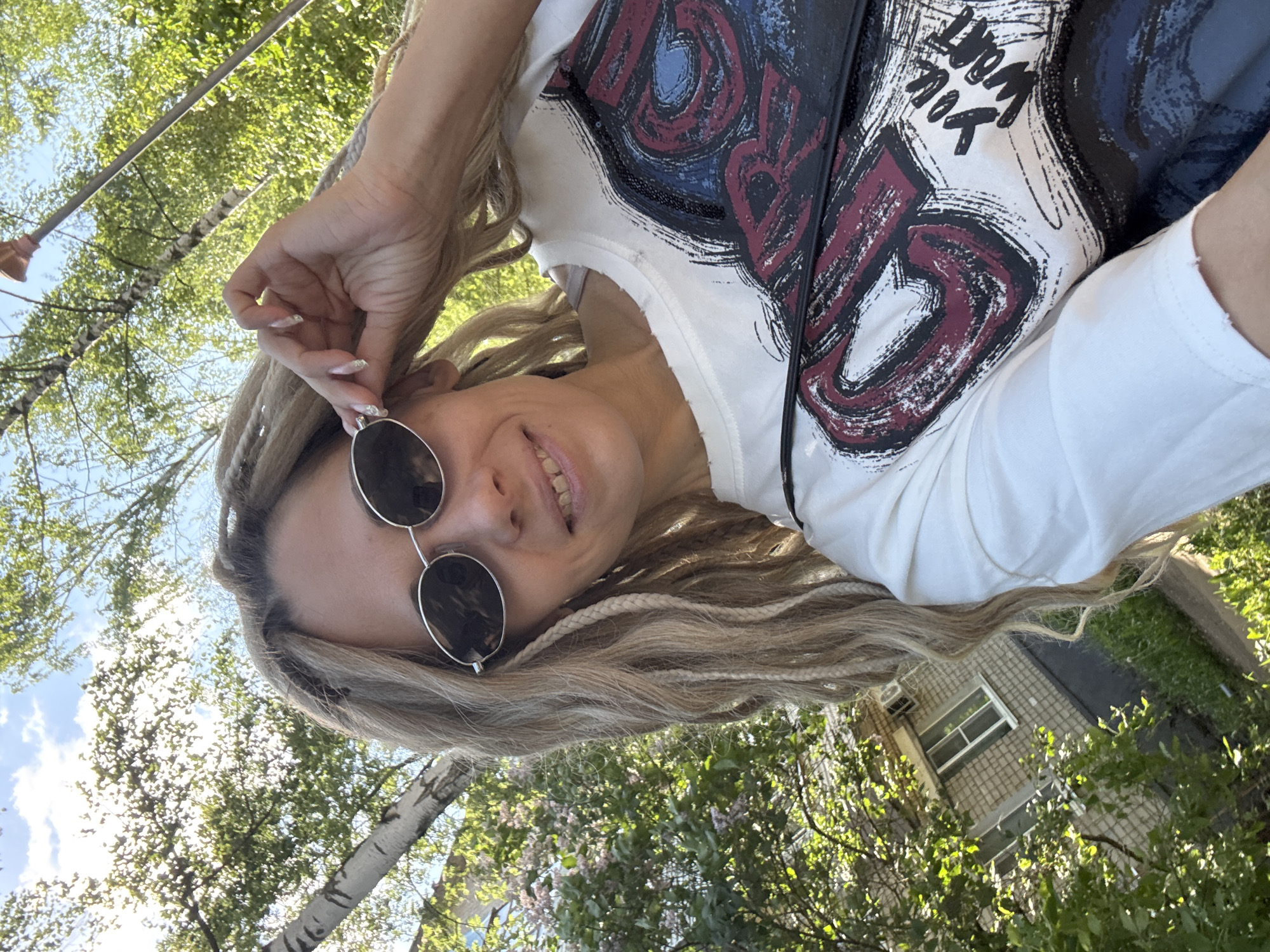 A woman with long blonde hair is wearing round sunglasses and a white t-shirt with a graphic design. She is standing outside, possibly in the city of Rusanovo, Russia. In the background, there are trees, a birch tree, and a building with a window. She has her hand near her ear and is smiling. The photo is taken from a low angle, looking up at her.