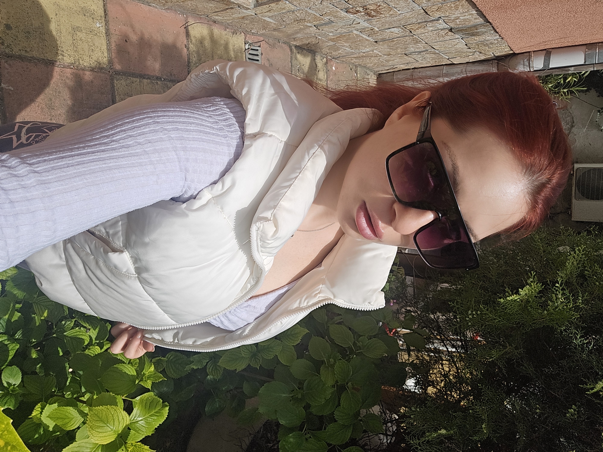 A woman with reddish-brown hair and sunglasses is positioned outdoors, presumably in Burgas, Bulgaria. She is wearing a light purple top and a white puffer vest, partly over a bed of green foliage. The ground is paved with brickwork. In the background, there are greenery and building elements including an air conditioning unit.