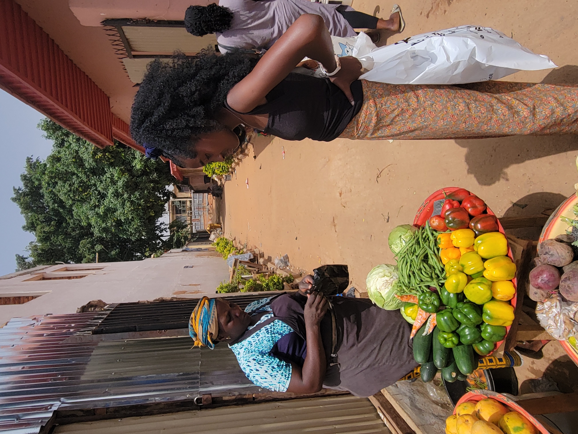 In Jos, Nigeria, a street scene unfolds with a woman examining produce from a vendor. The customer, wearing a black top and patterned skirt, looks into a white plastic bag. The vendor, in a blue and green patterned top and brown apron, is positioned behind a display of vegetables including bell peppers, tomatoes, green beans, cabbage, zucchini, and beets. A mango display is visible below the main vegetable arrangement. The background includes a dirt road, buildings with corrugated metal and partially damaged roofs, and a tree. Another person is walking away in the background.