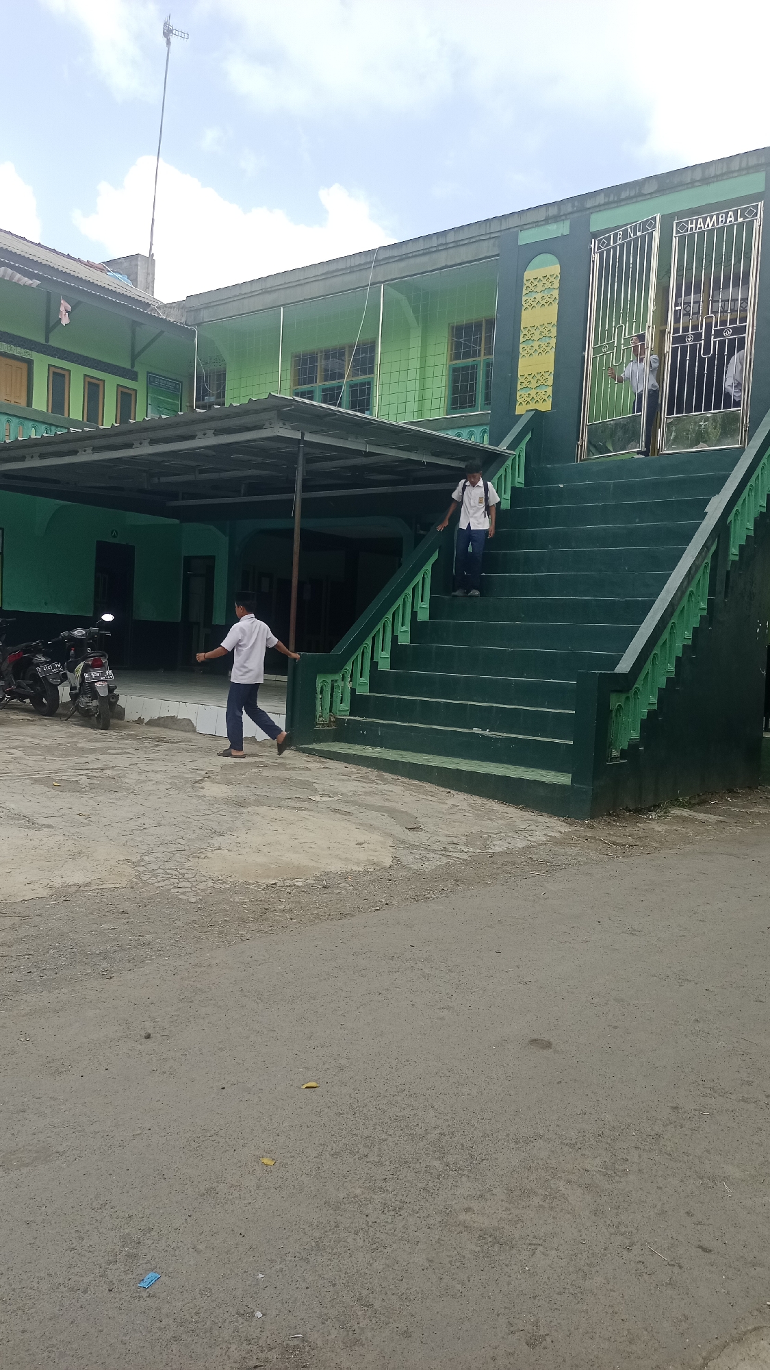 This image shows a building with a green exterior, possibly a school or religious institution, located in Karyamandala, Indonesia. The building has large windows and a green staircase leading up to an entrance with metal gates. Two young males, likely students based on their uniforms, are present in the scene. One is walking down the stairs, and the other is on the ground level, walking away from the stairs. Several motorcycles are parked in front of the building. The ground is paved with a mixture of asphalt and dirt. The sky is partly cloudy.