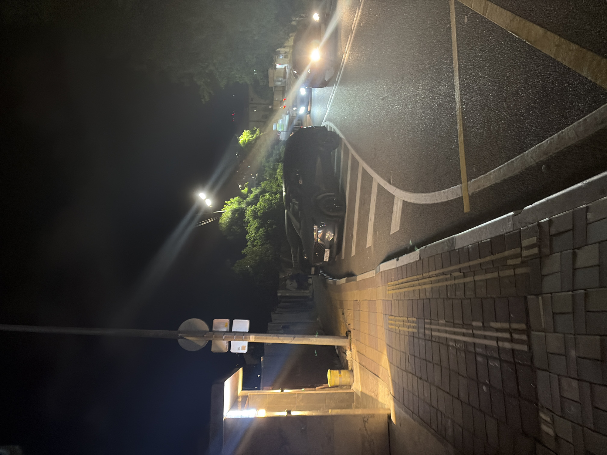 This image depicts a night scene on a street in Sochi, Russia. A dark SUV is parked on the right side of the road. Further down the road, two other vehicles are visible, their headlights illuminating the dark. A sidewalk runs along the left side of the road, paved with brick-like tiles. A yellow trash bin is situated on the sidewalk near a structure with a lit overhang. A traffic light pole with two rectangular signs is also present on the sidewalk. The road has visible lane markings, and trees and foliage line the sides of the street. The overall atmosphere is dimly lit, with artificial light sources casting shadows and highlighting certain areas.