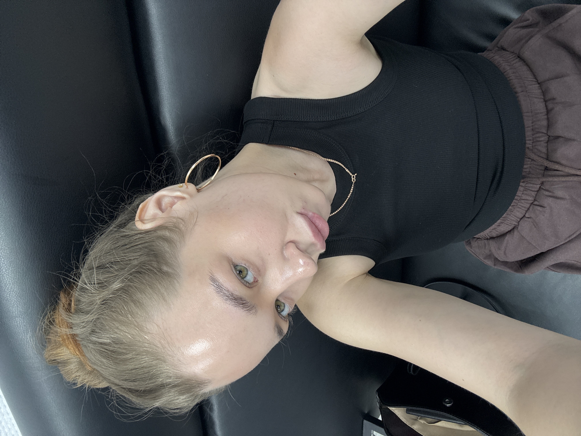 A young woman is reclining on a black leather surface. She wears a black ribbed tank top and brown drawstring pants. Her blonde hair is pulled back into a bun. She is wearing a gold hoop earring and a thin gold necklace. The location is potentially Chaikovsky, Russia.