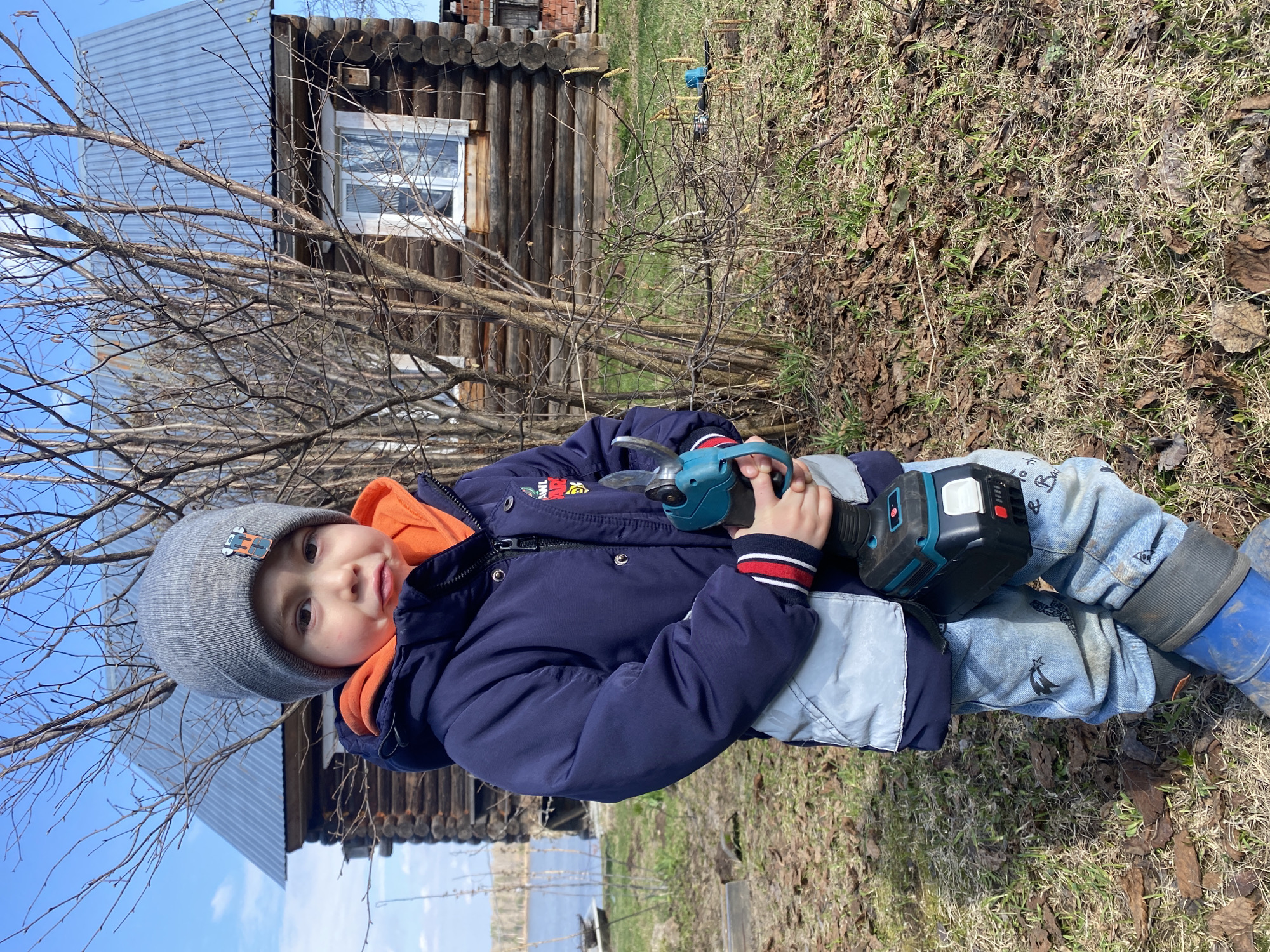 A young child in a gray hat, orange hoodie, and blue jacket is holding a teal and black cordless pruning shear in their hands. The child is wearing light blue jeans and blue rubber boots. They are standing in a grassy area with dead leaves and bare trees. Behind the child is a wooden log cabin with a corrugated metal roof. The location is Ольховка, Russia.