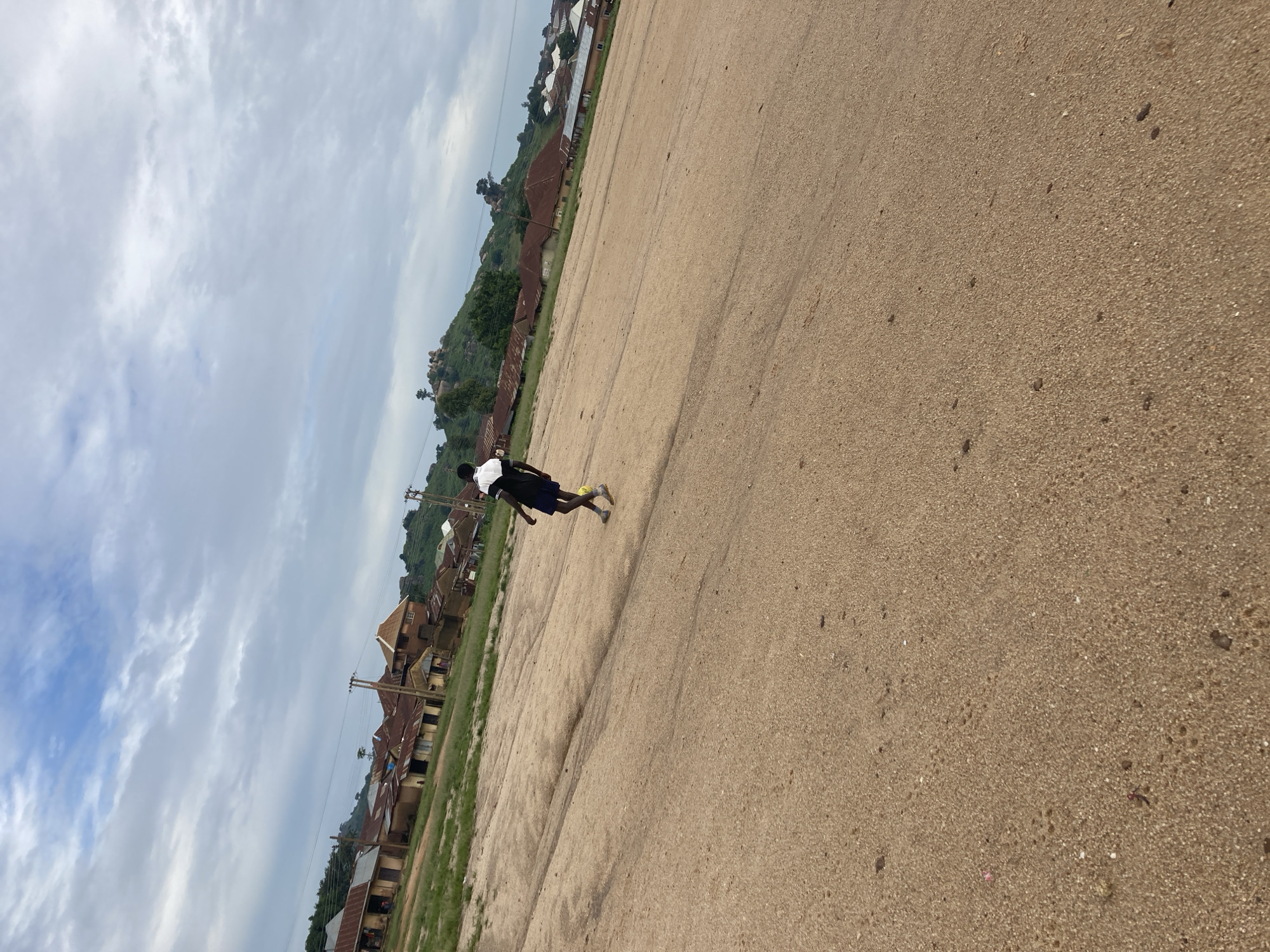 A young man is walking across a large, sandy, outdoor area. He is wearing a black and white shirt and blue shorts, and he has a yellow ball at his feet. The sandy area is in the foreground, and behind it, there is a slight incline with some green grass and a scattering of buildings. The buildings are mostly single-story structures with corrugated metal roofs, and they appear to be residential. In the background, there is a rocky hillside covered in green vegetation. The sky is cloudy and overcast. The scene is consistent with an outdoor area in a town or village in Jos, Nigeria, likely a dusty or unpaved area used for recreation or as a pathway.