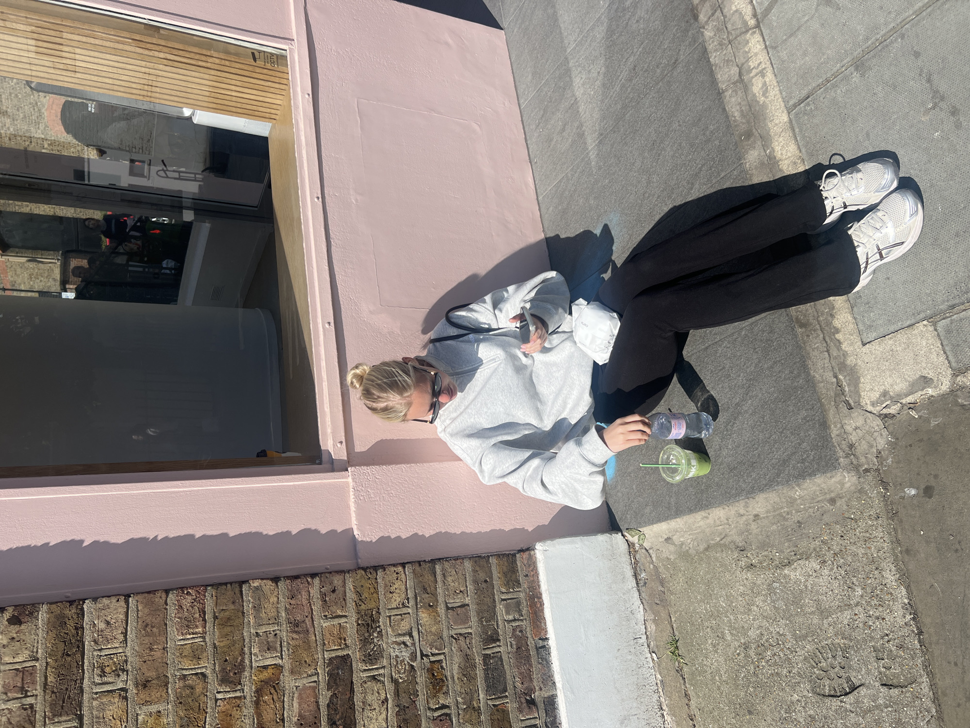 A young woman sits on the pavement next to a pink-painted building, possibly in London, United Kingdom. She is wearing a light grey sweatshirt, black trousers, and white trainers. Her blonde hair is tied up in a bun. She is holding a plastic water bottle and a green smoothie or drink in a plastic cup. The building next to her has a window with a wooden blind. The pavement is cracked and worn. The lower portion of the building is brick.