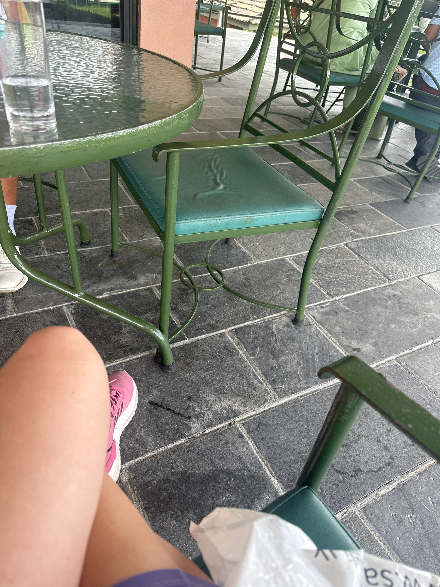 The image shows a close-up of a glass of water on a round green metal table, with green metal chairs and a tiled floor. In the foreground, a person's leg and pink sneaker are visible. In the background, other people are seated at tables. The setting appears to be an outdoor cafe or restaurant. The location is Kageshwori Manohara, Nepal.
