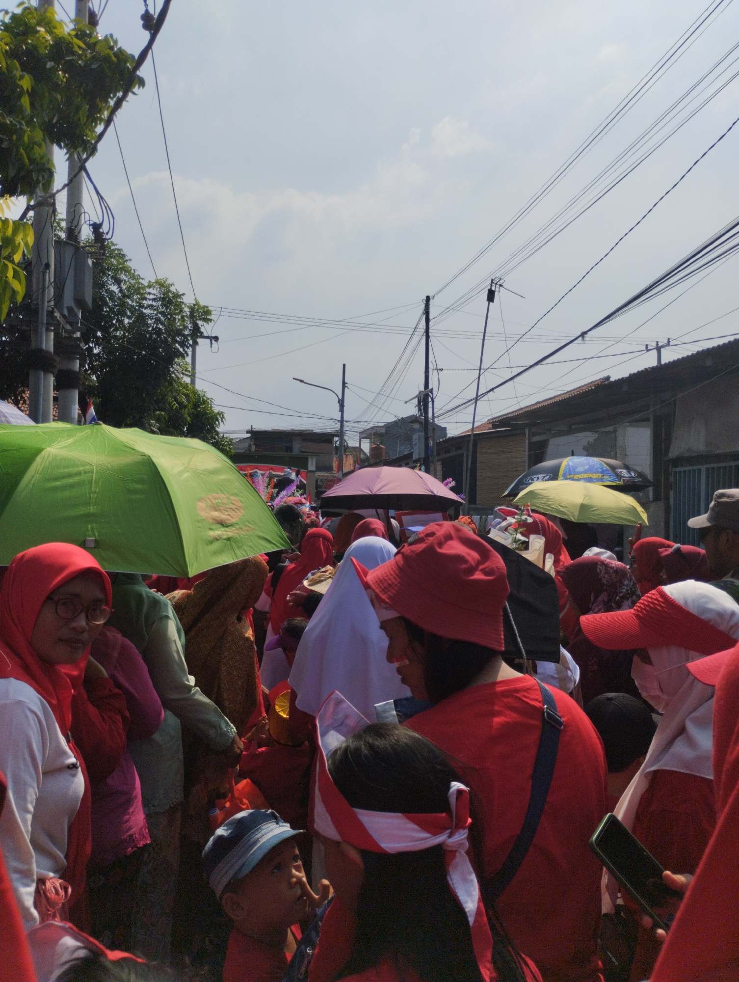 The image captures a crowded outdoor scene, likely a public event or gathering, under a bright sky with scattered clouds. A prominent green umbrella shields a portion of the crowd. Many individuals are wearing red clothing and headwear, with some also adorned in white and red fabric bands, suggesting a patriotic or celebratory theme. Umbrellas of various colors, including pink and yellow, are also visible, offering shade. Power lines crisscross the sky, supported by utility poles, indicating an urban or semi-urban setting. Buildings with tiled roofs are visible in the background, hinting at residential or commercial structures. The presence of a young child in the foreground, wearing a blue and white striped hat and looking upwards, adds to the sense of a community event. The overall atmosphere appears to be one of congregation and shared activity. The location is identified as Bekasi, Indonesia.