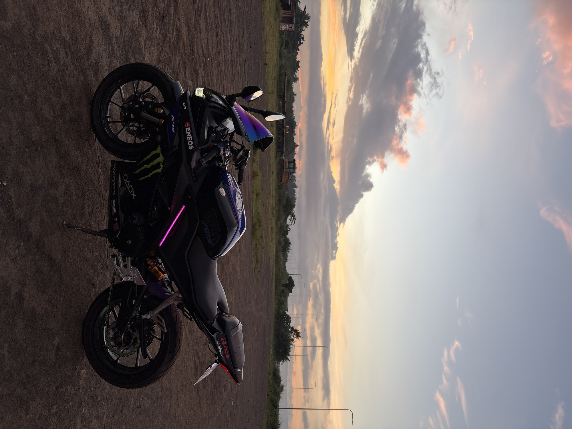 A black Yamaha R15 motorcycle is parked on a dirt lot under a dusky sky with pink and orange clouds. The motorcycle has a custom purple windshield and neon pink underglow lights. In the background, there are several lampposts, trees, and some construction equipment. The location is identified as Davao City, Philippines.