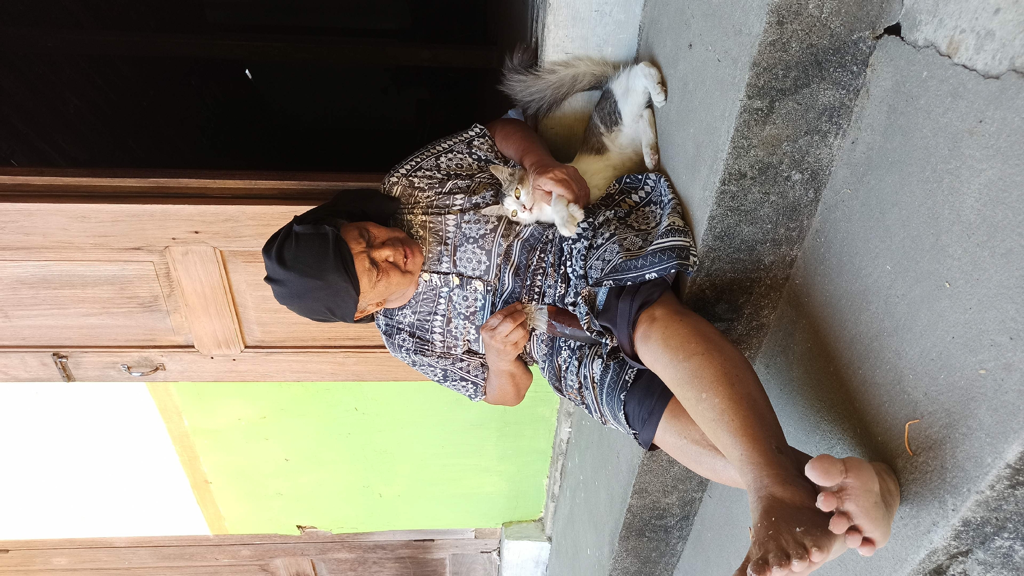 An elderly Indonesian woman, wearing a black headscarf and a patterned dress, sits on concrete steps outside a building with a wooden door. She holds a small bottle with dark liquid in her left hand and strokes a white and grey cat nestled in her lap with her right. The cat, with green eyes, looks upwards at the woman. Behind the woman, a bright green wall is visible to the left, and the wooden door is open to a dark interior on the right. The scene appears to be in the city of Simo, Indonesia, given the architectural style and the woman's attire.