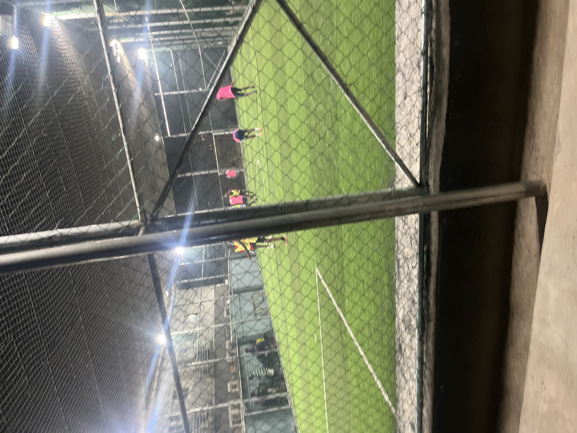 The image shows a fenced sports field, likely for soccer, with several people playing on the green artificial turf. The view is taken from behind a chain-link fence. The setting appears to be an indoor or covered facility with lights visible. The precise location within the city of Unknown, Unknown, cannot be determined from the image.