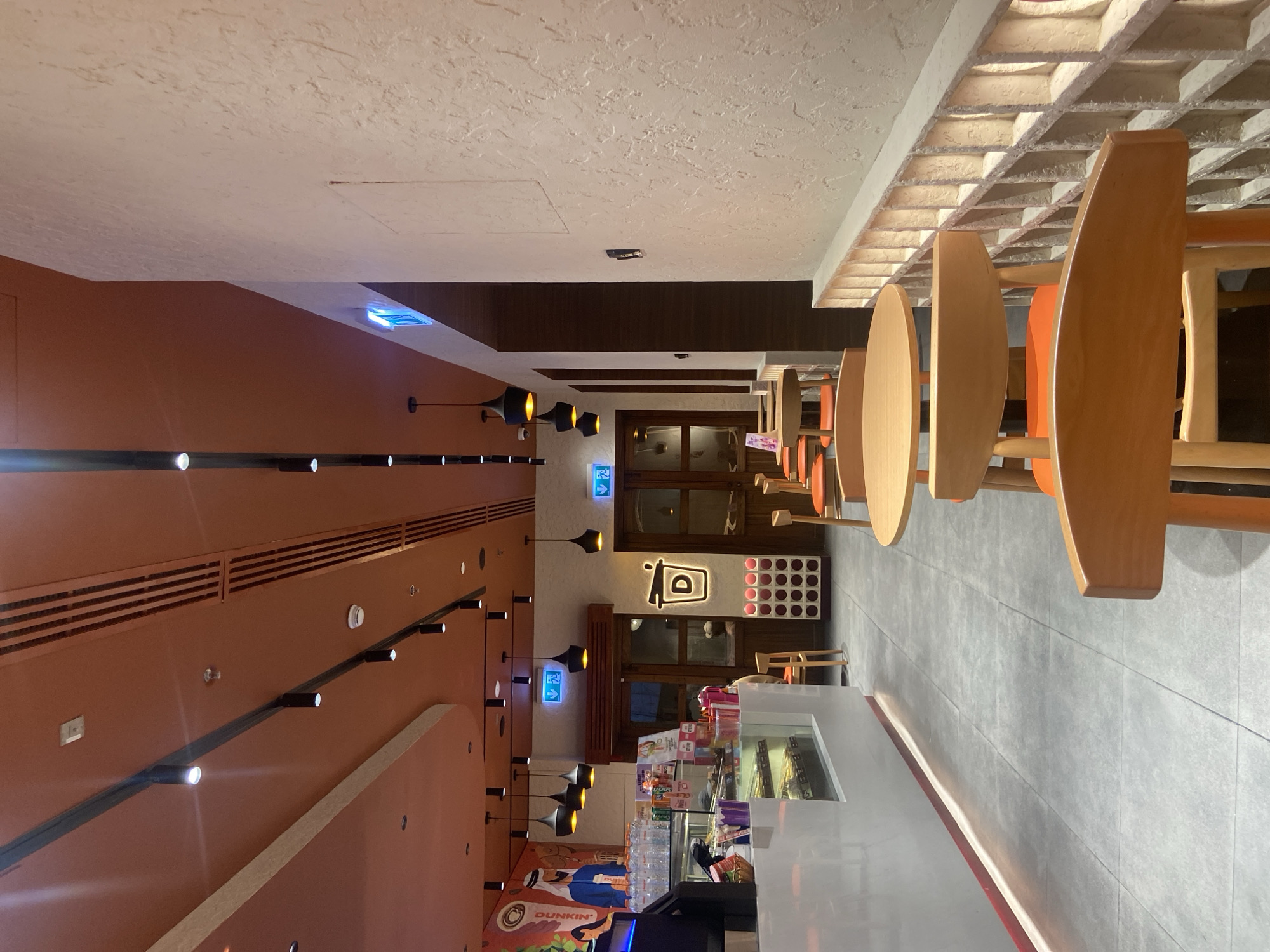 The image depicts the interior of a Dunkin' Donuts cafe in Deira, United Arab Emirates. The cafe features an orange ceiling with track lighting and decorative wooden slats. The walls are textured and light-colored, with a decorative brick partition along the right side of the image.
In the foreground, several wooden chairs with orange upholstery surround light-colored wooden tables. A counter area is visible on the left, with a display case showcasing donuts and other pastries. Behind the counter, a large mural depicts figures in traditional Middle Eastern attire.
Further back, wooden doors lead to other areas of the cafe. Blue exit signs with arrows are mounted above these doorways. The overall ambiance is modern and inviting, with a clear emphasis on the brand's signature colors and design elements. The scene is captured from a low angle, emphasizing the architectural details and the arrangement of the seating area. There are no visible people in the image.