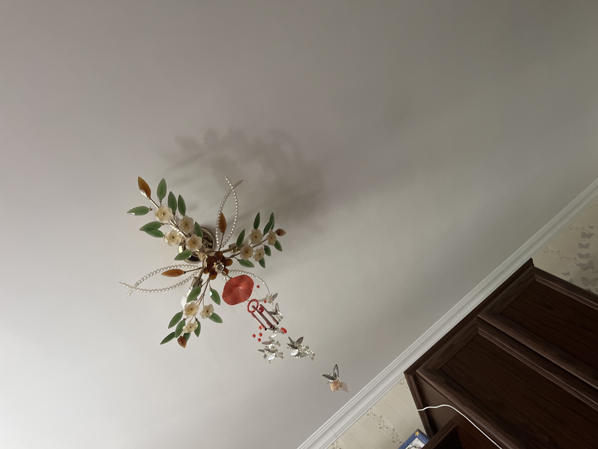 The image shows a decorative ceiling fixture with floral elements and several small, silver-colored hummingbird-like ornaments hanging from it. Some of the hummingbirds are positioned in flight, with iridescent wings. One hummingbird is closer to the camera and appears to be holding a small, light-pink object. A small red umbrella is also suspended from the fixture. The fixture itself is mounted on a white ceiling and is adorned with what appear to be pearl beads and amber-colored stones. Below the ceiling, a portion of a dark wood cabinet with ornate detailing is visible, along with a section of wallpaper with a subtle floral pattern. The lighting in the room is ambient, casting soft shadows on the ceiling. The photograph appears to have been taken from a low angle, looking upwards.