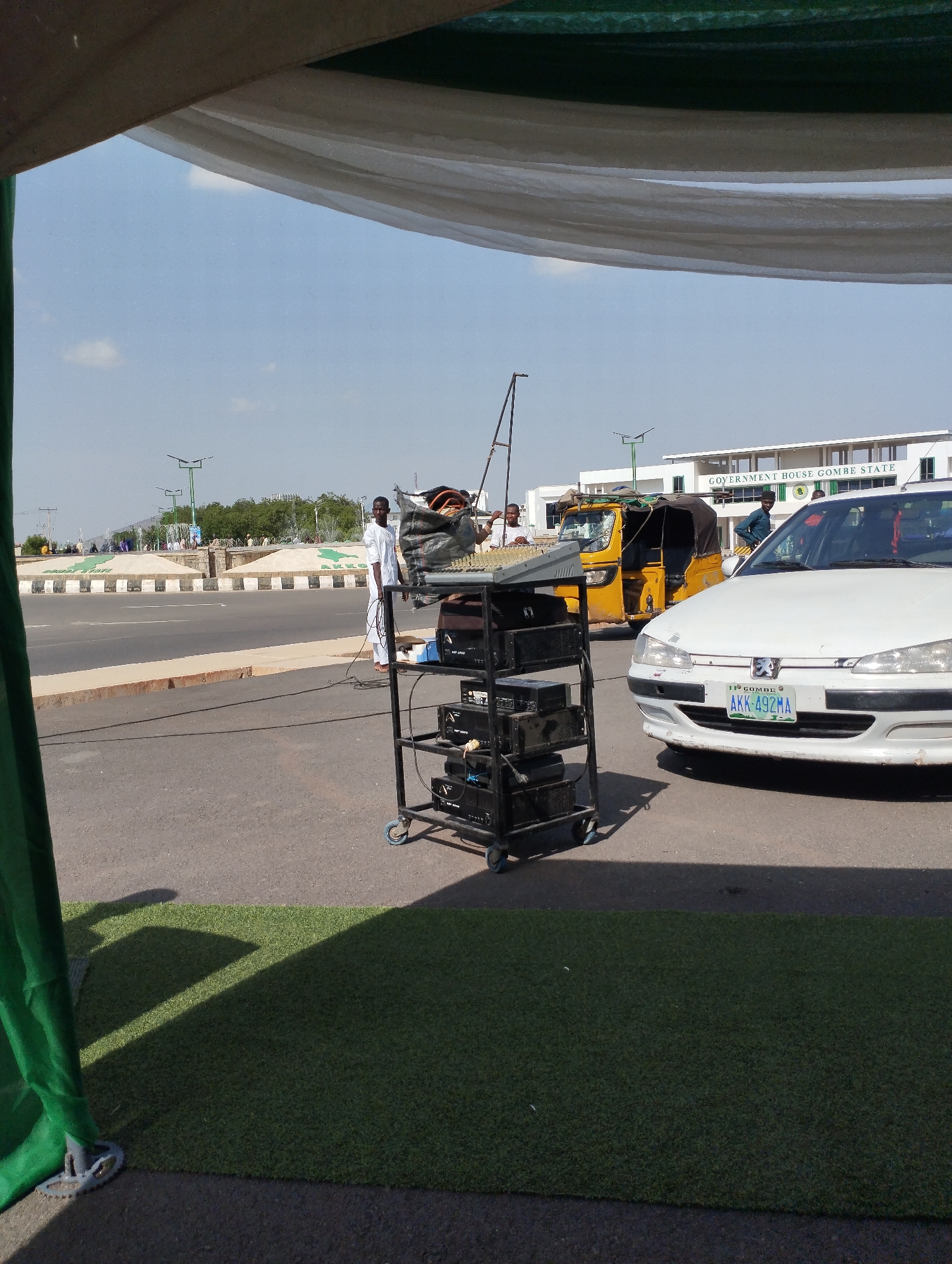 The image depicts a street scene in Gombe, Nigeria, in front of the "Government House Gombe State" building. A white Peugeot car with license plate "AKK 492MA" is parked on the road, next to a yellow auto rickshaw (tuk-tuk). A metal rack containing audio equipment is positioned on the street. Several people are visible in the background, including one man in a white outfit standing near the rack. A section of artificial green turf is visible in the foreground, likely part of a temporary setup. A white and green fabric structure frames the upper part of the image.