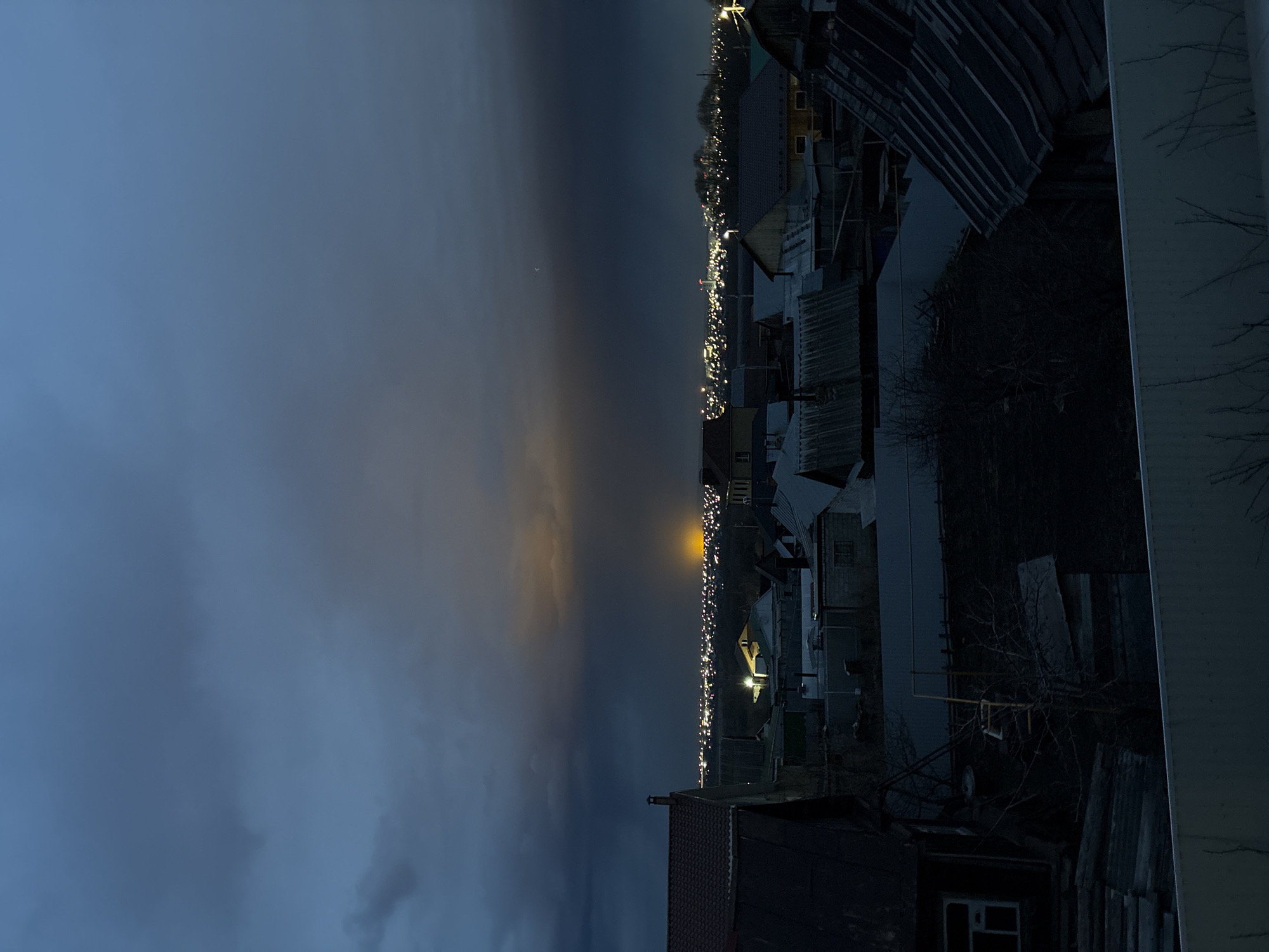 The image depicts a nighttime view of a residential area in Ulyanovsk, Russia. The sky is dark and overcast with a slight glow on the horizon. Below the sky, a street is visible with numerous lights from houses and streetlamps. The houses are mostly small, single-family dwellings with metal roofs. Some trees are visible in the foreground. A window and a chimney of a nearby house is visible at the bottom. The image is slightly rotated.