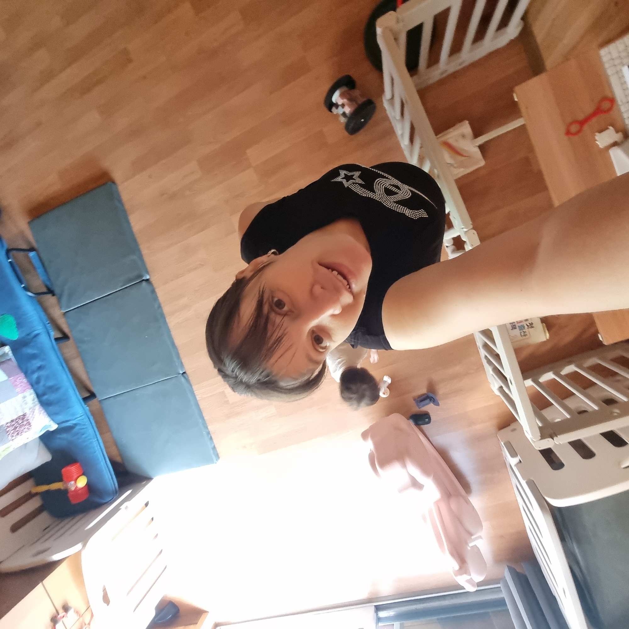 The image shows a woman taking a selfie from a high angle, looking up at the camera. She is wearing a black t-shirt with a white design on it. In the background, a baby is crawling on a wooden floor. There are several objects in the scene, including a white crib, a blue couch with pillows, and several toys, one of which appears to be a red toy hammer. The environment appears to be a domestic indoor setting, likely a child's play area or bedroom, with wooden flooring and a white crib. The lighting suggests it is daytime, with natural light coming in from the left side of the frame. There is no visible indication of the location being in Dunnae-myeon, South Korea, or any other specific geographical location.