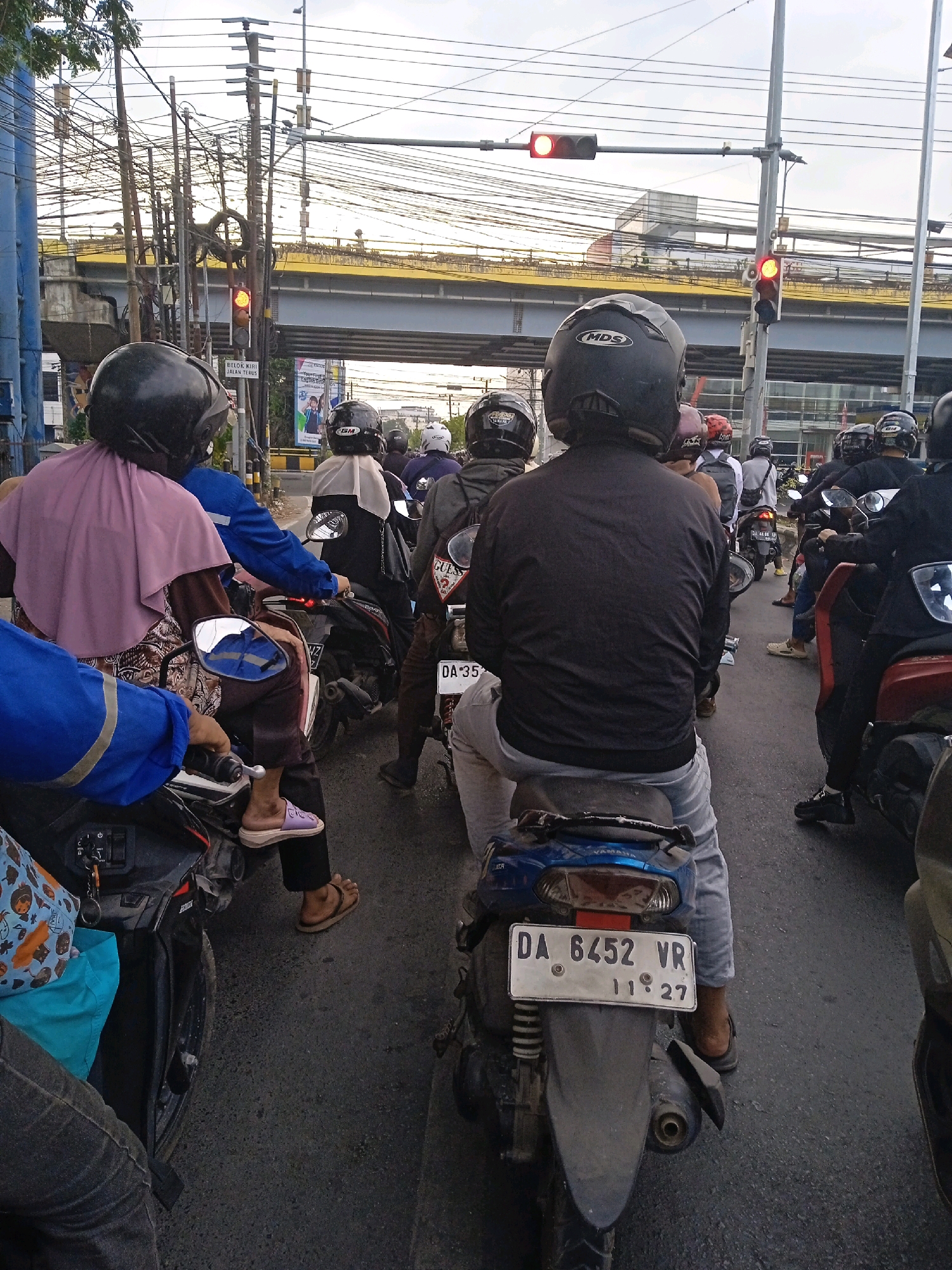 This is a street-level, eye-level medium shot of a traffic jam in Banjarmasin, Indonesia. The foreground features the backs of several motorcyclists wearing helmets, stopped at a traffic light. The motorcyclist in the center is wearing a black jacket and grey pants, with a blue Yamaha scooter. The license plate reads "DA 6452 VR" and "11 27". To the left, a motorcyclist in a pink hijab and blue jacket is visible. In the background, a yellow overpass with a traffic light showing amber is visible. Overhead, a complex network of power lines crisscrosses the sky. Buildings and trees are also visible in the distance. The scene is consistent with a typical urban environment in a Southeast Asian city.