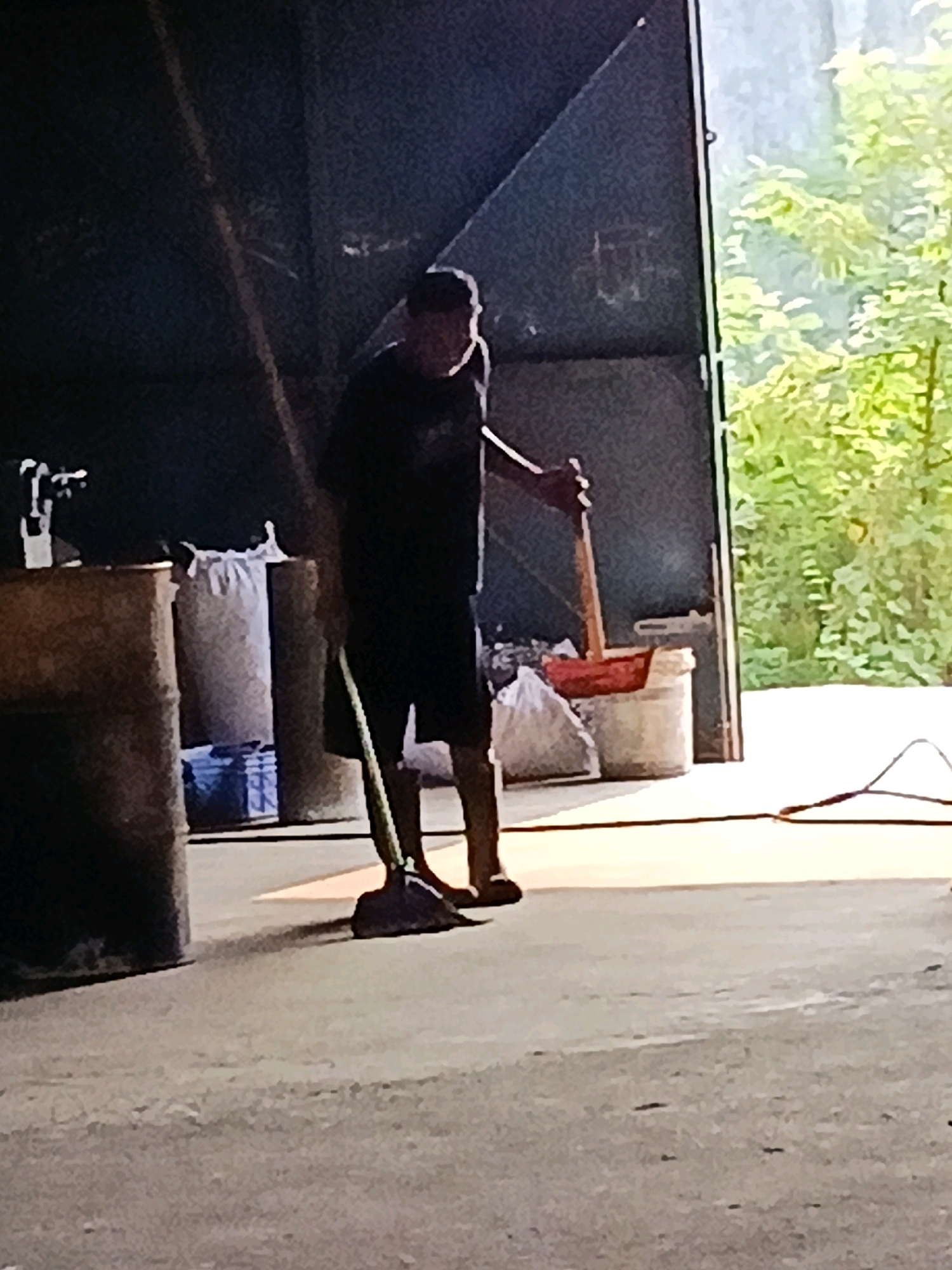 A person is sweeping the ground in front of an open doorway, which leads to an outdoor area with trees. The person is wearing a dark t-shirt, dark shorts, and a dark baseball cap. They are holding a broom and are in the process of sweeping. The ground is concrete. There are some large cylindrical objects and bags visible inside the doorway to the left. There is a bucket with a red object in it to the right of the person. The image appears to be taken in a storage area or a workshop.