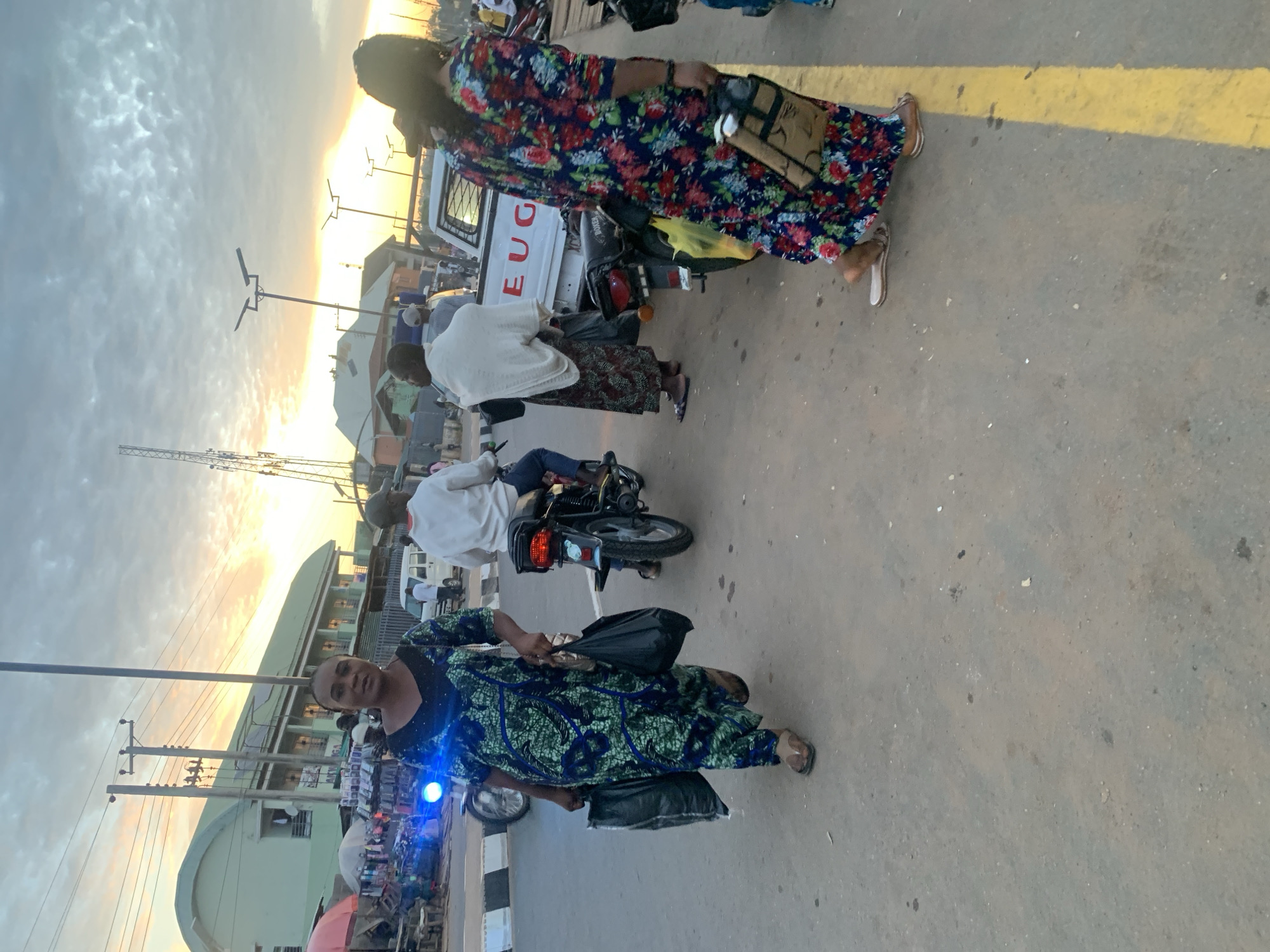 The image depicts a street scene in Akwanga, Nigeria, likely during the late afternoon or early evening as indicated by the warm, low sunlight casting long shadows. Several people are present, with a focus on two women in the foreground. The woman on the left is wearing a blue and green patterned dress and is looking towards the camera with a neutral expression. She is carrying two black bags. The woman on the right is wearing a colorful floral dress and is walking away from the camera. She is also carrying a bag.
In the background, other individuals are visible, some on motorcycles and others walking. A white truck with the partially visible text "EUG" on its side is parked nearby. The setting appears to be a busy street with various buildings and power lines visible. The ground is paved with asphalt, marked with a yellow line. The sky is overcast with some sunlight breaking through, suggesting a partly cloudy day. The overall atmosphere is one of everyday activity in a bustling urban environment.
