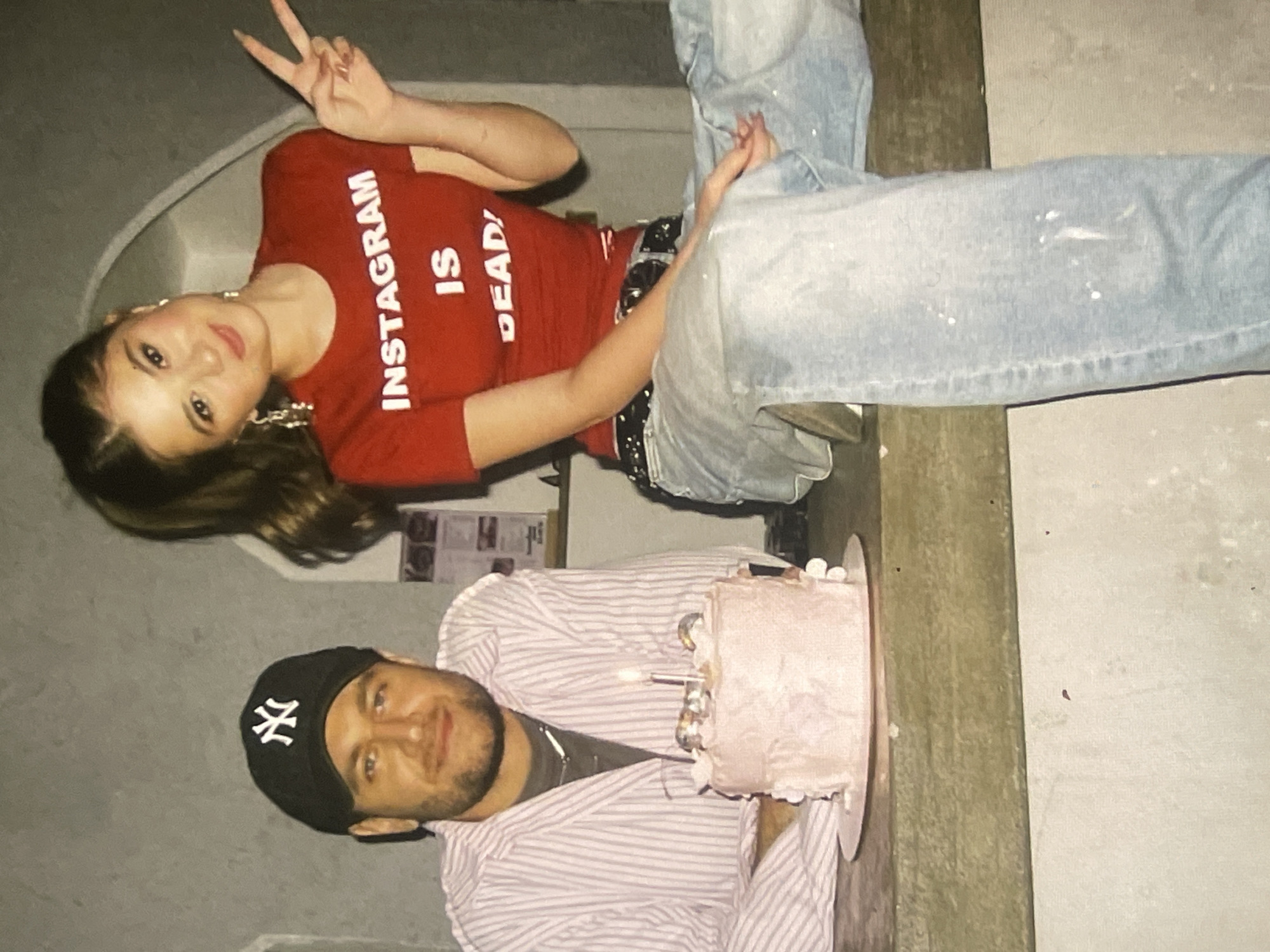 An indoor scene captures a celebratory moment featuring a man and a woman. The man, positioned on the right side of the frame, wears a black New York Yankees baseball cap, a light-colored striped shirt over a dark t-shirt, and has a beard. He is smiling and looks towards the viewer. In front of him, resting on a counter or table, is a light pink frosted cake with several lit candles, indicative of a birthday celebration.
To the left of the frame, a woman with long, dark hair leans or sits on the same surface, smiling and making a peace sign with her right hand. She is dressed in a red long-sleeved t-shirt with the bold white text "INSTAGRAM IS DEAD!" across the chest. She also wears light blue jeans, which appear to have white paint splatters, and a dark belt. Both individuals appear happy and relaxed. The background is simple, featuring light-colored walls and a wooden beam or counter that runs horizontally across the image, separating the two subjects. The event is taking place in Poltava, Ukraine.