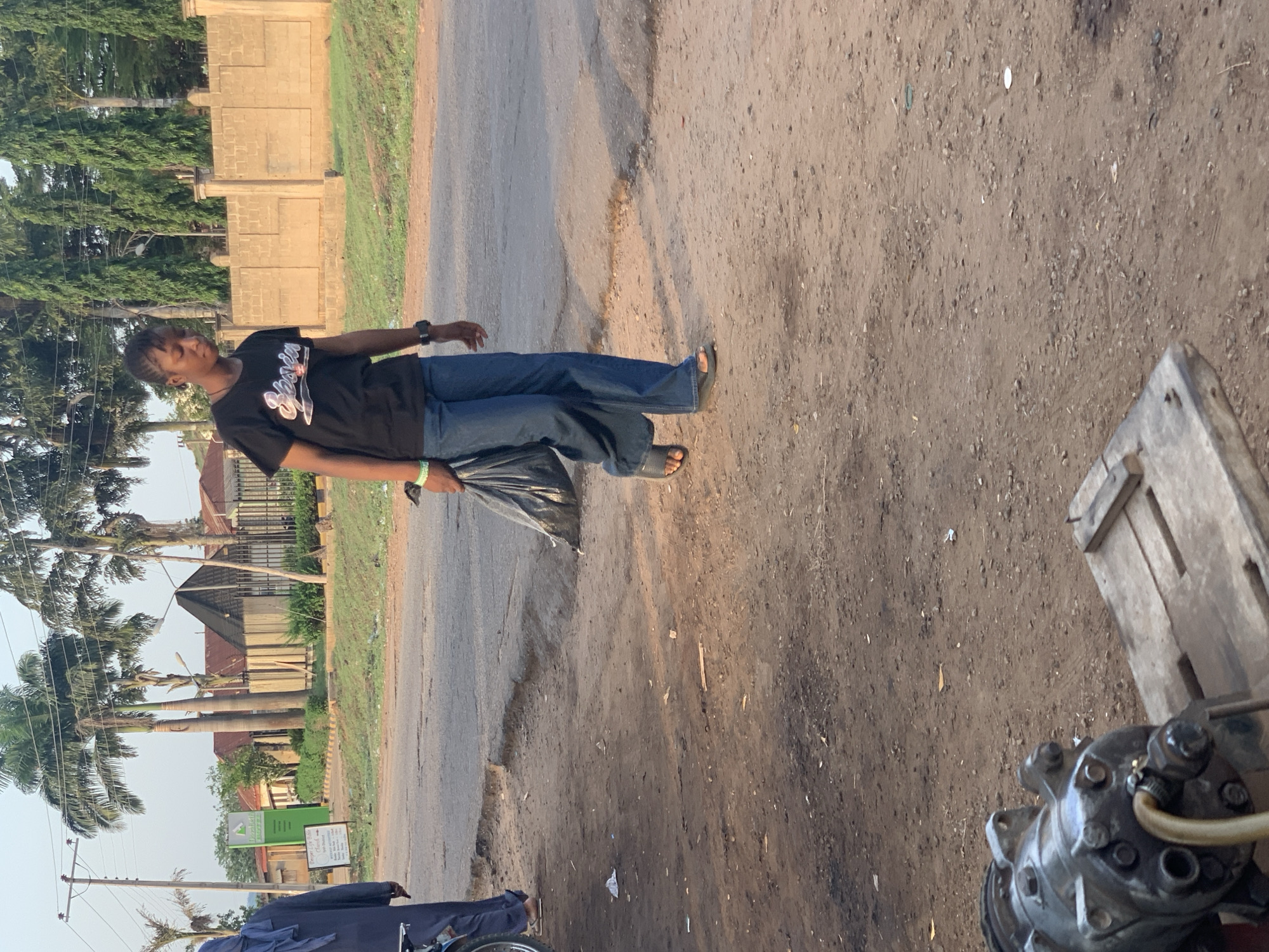 The image shows a person walking on a street in Gbeleko, Nigeria. The person is wearing a black t-shirt, blue jeans, and sandals, and is carrying a black bag. The street is paved, but with some broken sections, and there is dirt and gravel on the side of the road. There are buildings and trees in the background. A vehicle with a blue cover is visible in the lower left, and a machine of some kind is in the lower right.