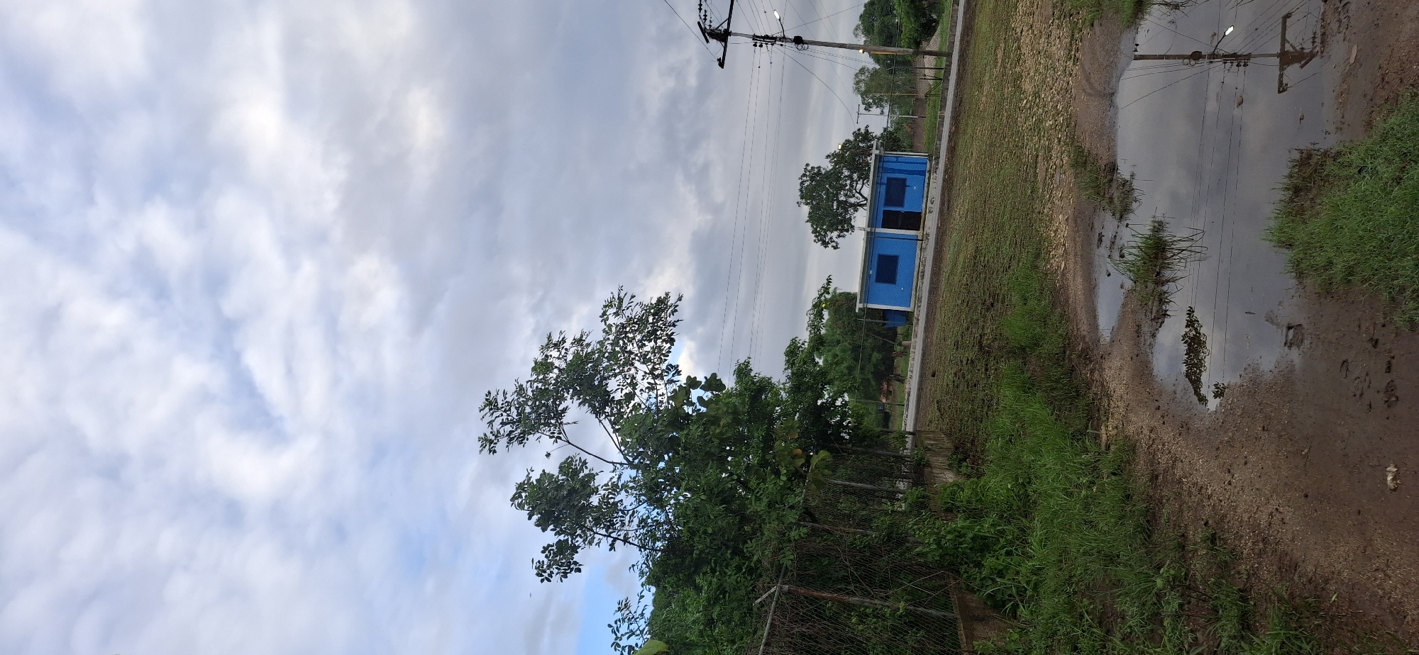 The image depicts an outdoor scene with a cloudy sky. In the middle ground, a bright blue modular building with dark window frames is visible, situated next to a dirt path. A utility pole with wires runs along the side of the path, extending towards the building. The foreground is dominated by lush green grass and a chain-link fence. To the right of the path, there's a large puddle reflecting the sky and utility pole. The overall impression is that of a somewhat overcast day in a rural or industrial setting. The image's orientation is portrait, with the sky occupying the top portion and the ground elements below.