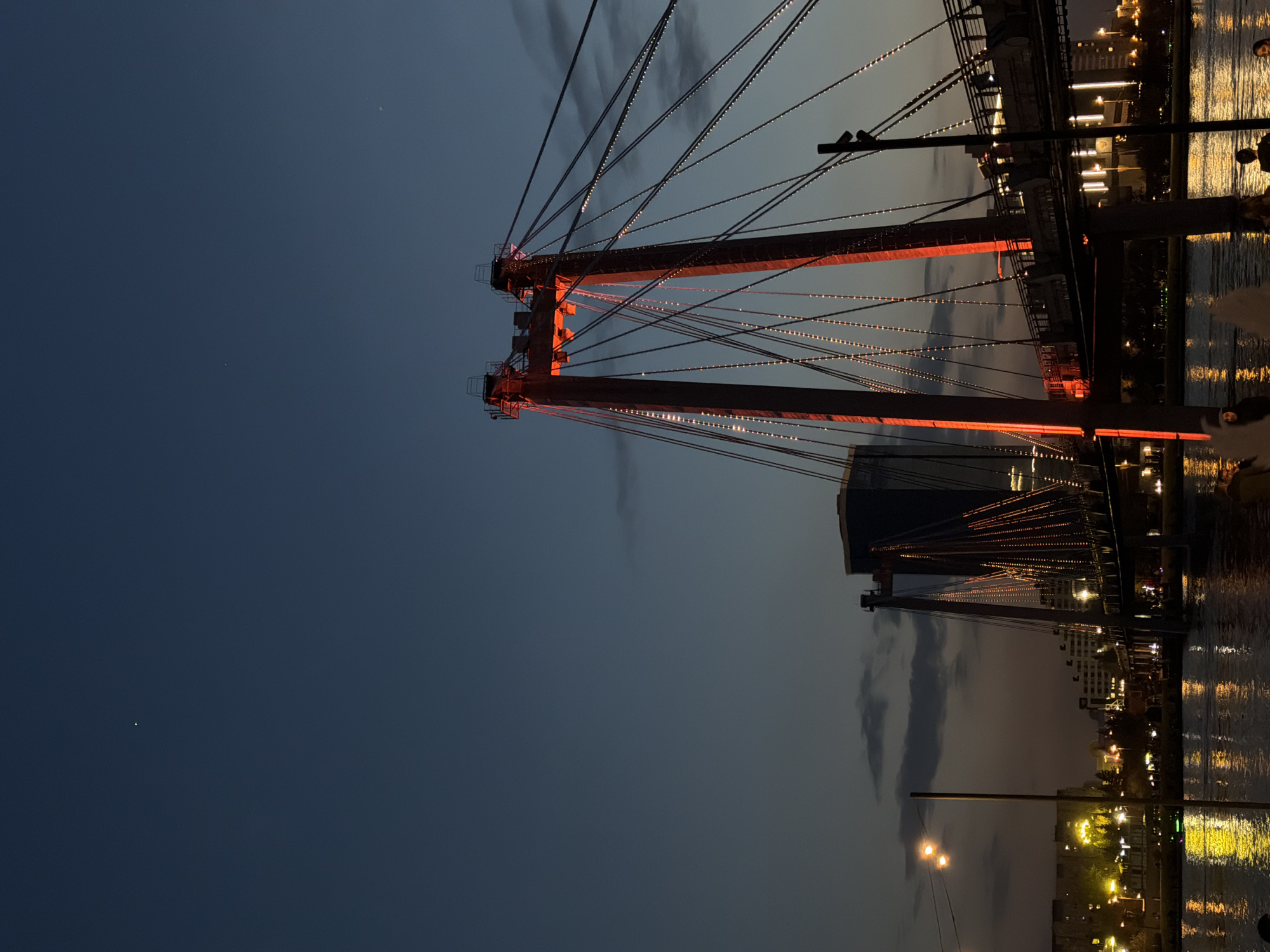 The image shows a nighttime view of a bridge in Krasnoyarsk, Russia. The bridge's supports are lit with orange lights, and lines of small lights run along its cables. In the background, city lights reflect on the water. The sky is dark with a few clouds.