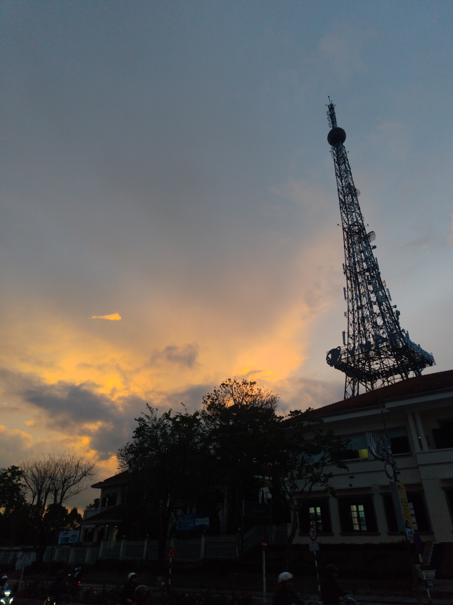 The image shows the Da Lat television tower at sunset in Da Lat, Vietnam. The tower is the tallest structure in the city. The sky is orange and gray. There is a large white building with a red roof under the tower. Trees are in front of the building. People on motorbikes are on the road.