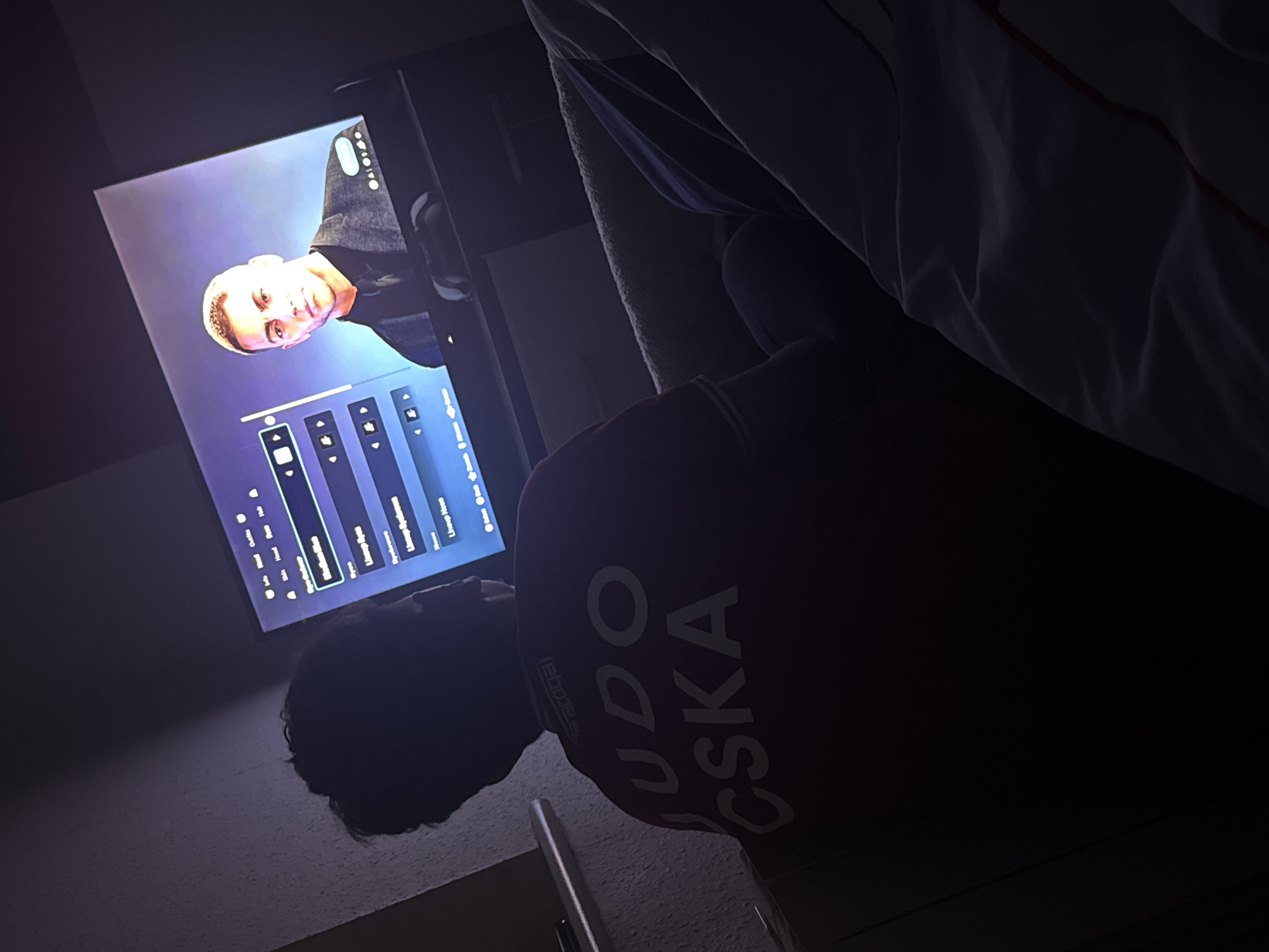 A person with short, dark hair is lying down and facing a television screen. The screen displays a man with short, blond hair, wearing a suit jacket. The television interface shows settings and options. The person lying down is wearing a red shirt with white lettering that reads "JUDO CSKA". The setting appears to be indoors, possibly a bedroom. There is no information to confirm the location as Welshpool, United Kingdom.