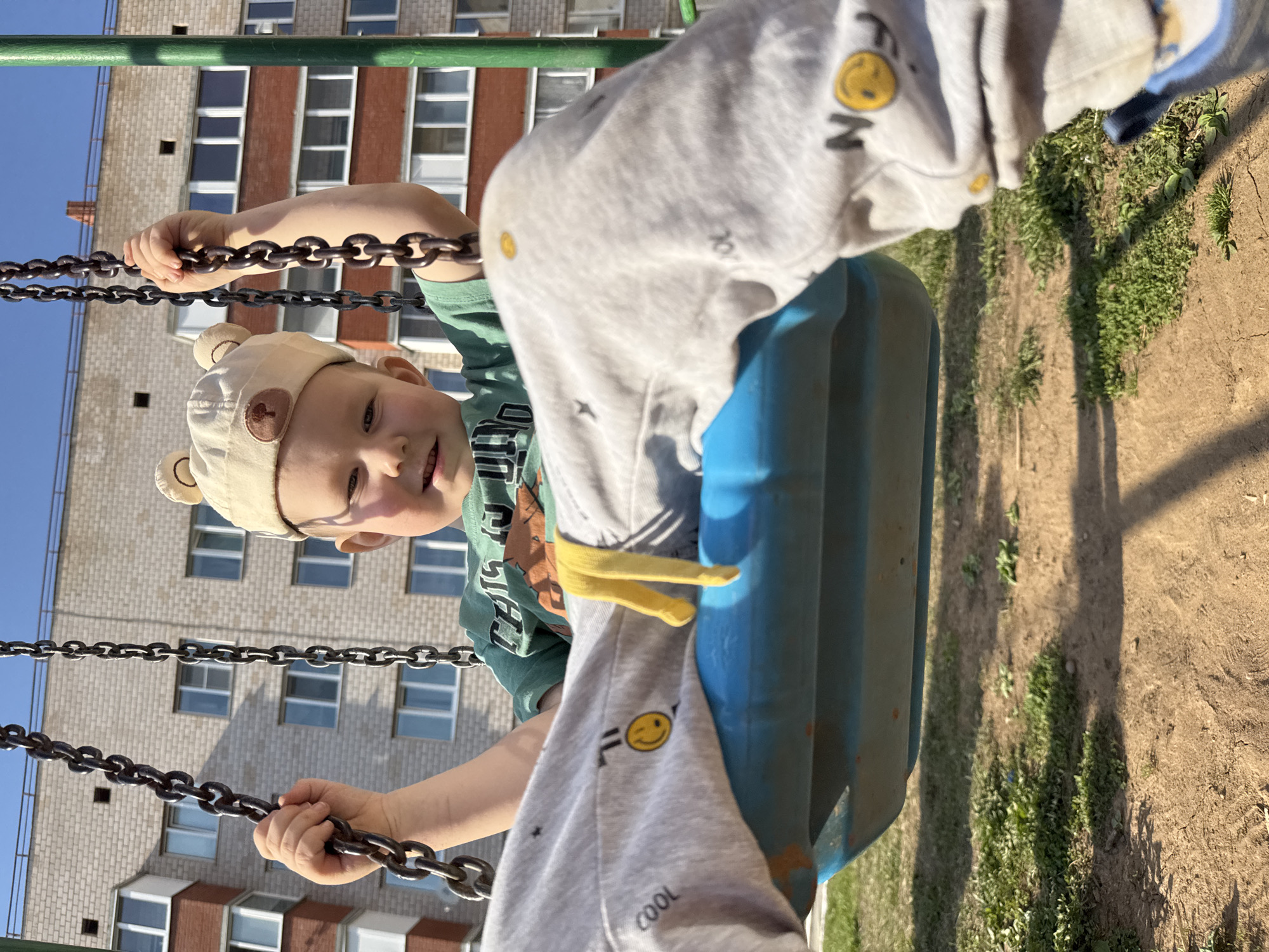 A young child wearing a beige hat with bear ears, a green t-shirt, and gray sweatpants is on a blue swing at a playground in Zav'yalovo, Russia. The child is holding onto the chains of the swing. Behind the swing, there is a multi-story brick building with windows. The ground beneath the swing appears to be a mix of dirt and sparse vegetation. The lighting suggests it is daytime.