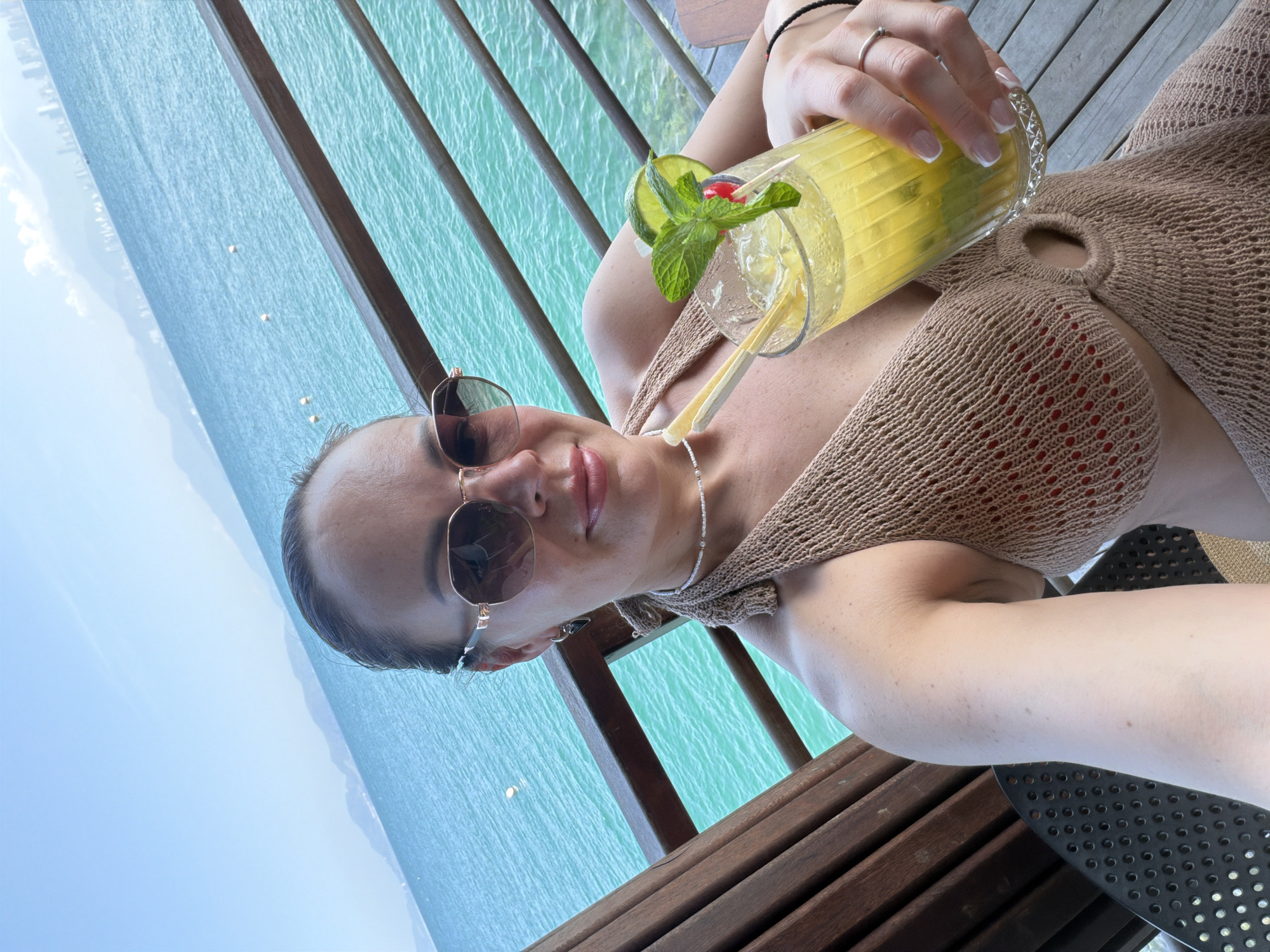 A young woman is captured in a selfie, relaxing in a tropical setting. She looks towards the camera with a relaxed expression, sporting sunglasses with gold frames and a hint of pink lipstick. Her hair appears to be pulled back, and she wears a delicate necklace and a silver ring. She is dressed in a light brown or beige knitted top, possibly part of swimwear, featuring a circular O-ring detail on the chest, with a glimpse of red fabric underneath.
She holds a tall glass cocktail, garnished with a lime wedge, fresh mint leaves, and a red cherry, complete with two straws. The drink is yellowish-green and filled with ice, suggesting a refreshing beverage.
The background reveals a stunning overwater scene, with vibrant turquoise ocean water visible through and beyond a wooden deck or railing. Distant mountains or land are faintly visible on the horizon under a clear blue sky, indicating bright, sunny daytime weather. The overall impression is one of leisure and vacation, likely at a resort in a beautiful coastal location such as Nha Trang, Vietnam. No text is visible in the image.