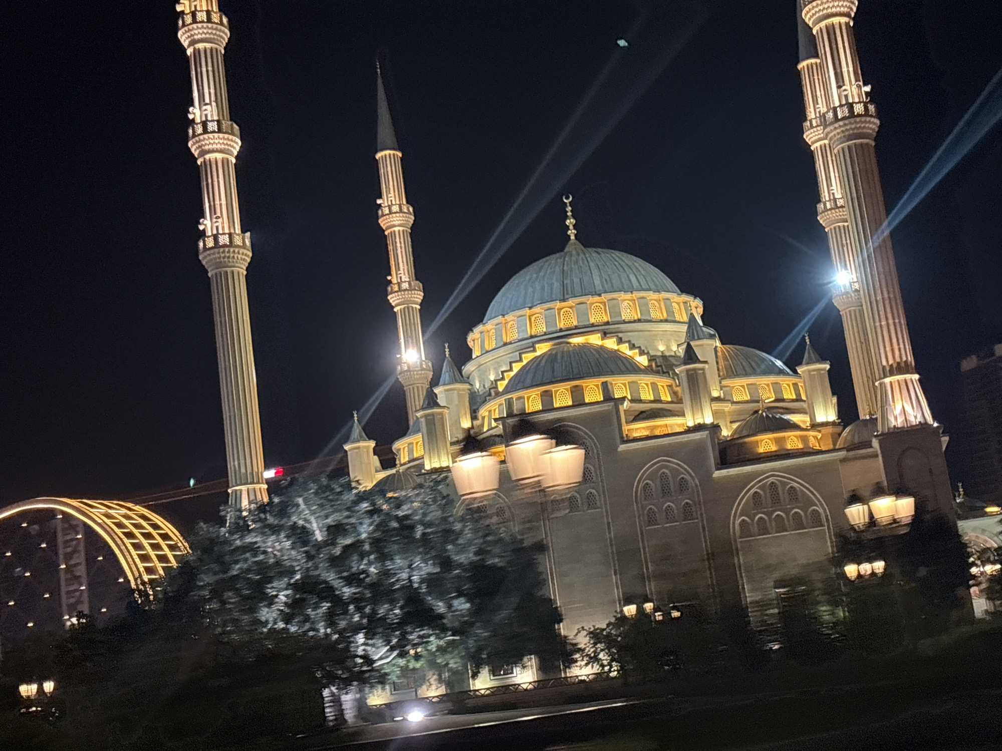 The image shows the Akhmad Kadyrov Mosque, also known as the "Heart of Chechnya," in Grozny, Russia, at night. The mosque is illuminated by spotlights, highlighting its white marble facade, large central dome, and four tall minarets. The minarets are intricately decorated with vertical ribbed patterns and balconies. The main dome is a light blue color and is surrounded by smaller domes and turrets. Numerous windows with arched openings are visible on the mosque's walls, also lit from within.
In the foreground, a large tree with dense foliage obscures part of the mosque's lower section. The tree appears to be in motion blur, suggesting a slight movement in the camera or wind. To the left of the tree, a section of a large, illuminated, arched structure with a lattice design is visible. In the background, the dark night sky is punctuated by white light beams emanating from the mosque or its surroundings, creating a dramatic effect. There are also streetlights visible in the lower part of the image. No people are clearly discernible in the scene. The overall impression is one of grandeur and architectural beauty, enhanced by the night lighting.
