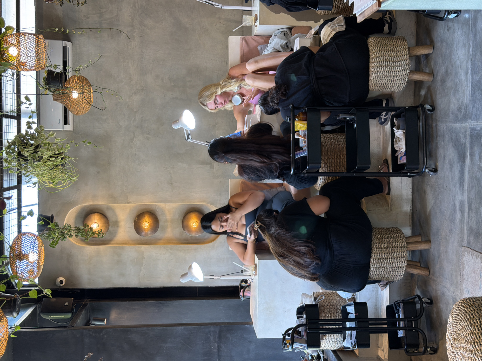 The media file shows a group of people in a brightly lit nail salon. The room has a modern, minimalist aesthetic with concrete walls and natural materials like rattan for the lampshades and stool covers.
There are at least three women visible, appearing to be clients and technicians. One woman with blonde hair is seated and having her nails done by another woman. A third woman with dark hair is also seated, seemingly waiting or engaged in her phone. The atmosphere appears relaxed and professional.
The salon has multiple workstations, each equipped with a desk lamp and tools. Plants are incorporated into the decor, adding a touch of greenery. The lighting suggests it is daytime, and the indoor setting indicates no specific weather conditions are relevant.
Notable details include the woven rattan elements, the concrete walls, and the sleek modern furniture. The focus is clearly on the nail services being provided.
The overall scene is one of a contemporary nail salon in North Kuta, Indonesia, where clients are receiving beauty treatments.