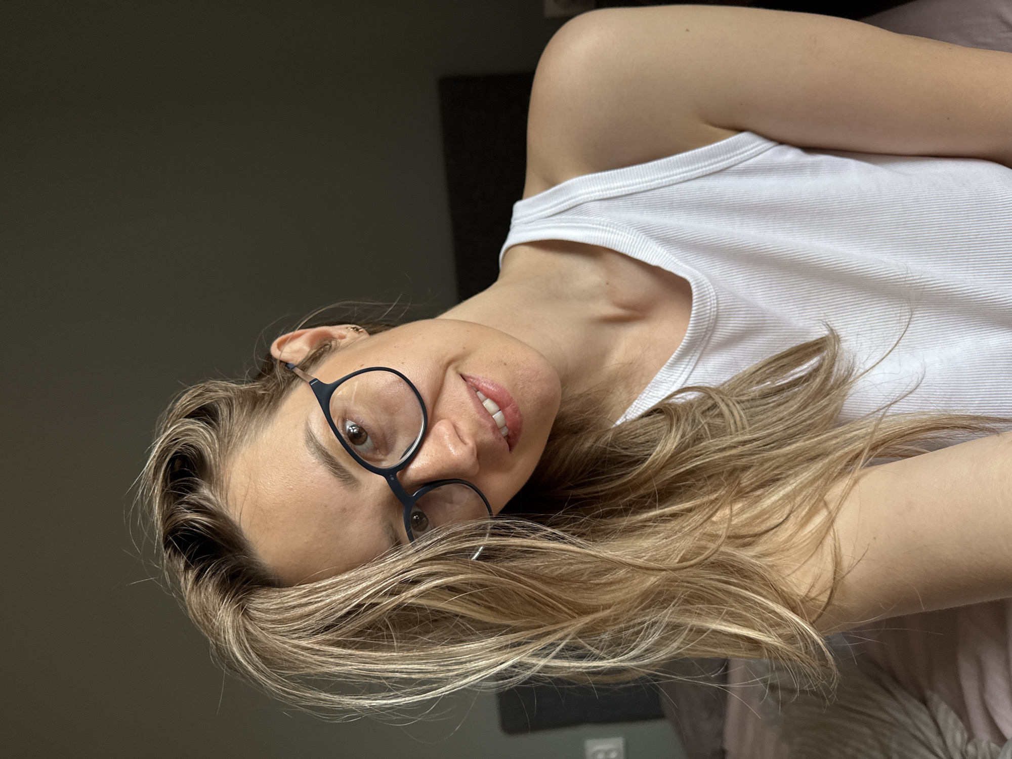 A woman wearing glasses and a white tank top is lying down and looking towards the viewer. Her blonde hair is spread around her. The background appears to be a room with dark grey walls. There is no indication that this image was taken in Chaikovsky, Russia.